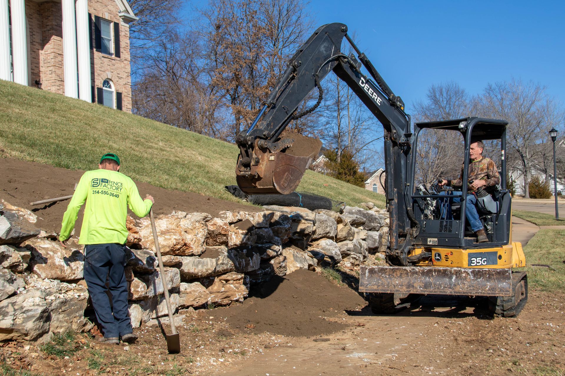 Man operating excavator, another man nearby, working on a stone retaining wall in front of a house.