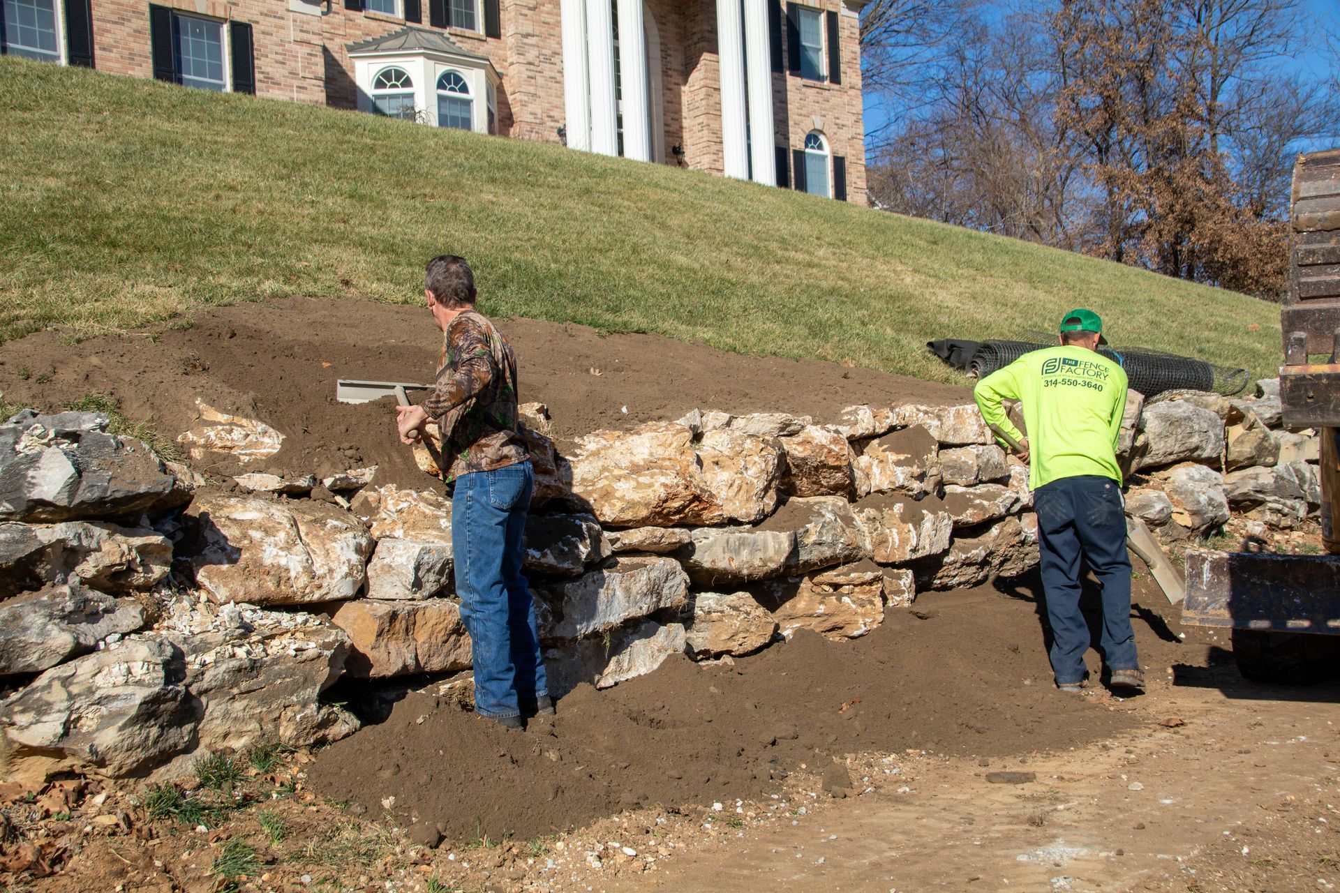 Two workers building a rock wall in front of a hillside home; dirt, rocks, blue jeans, and green shirts.