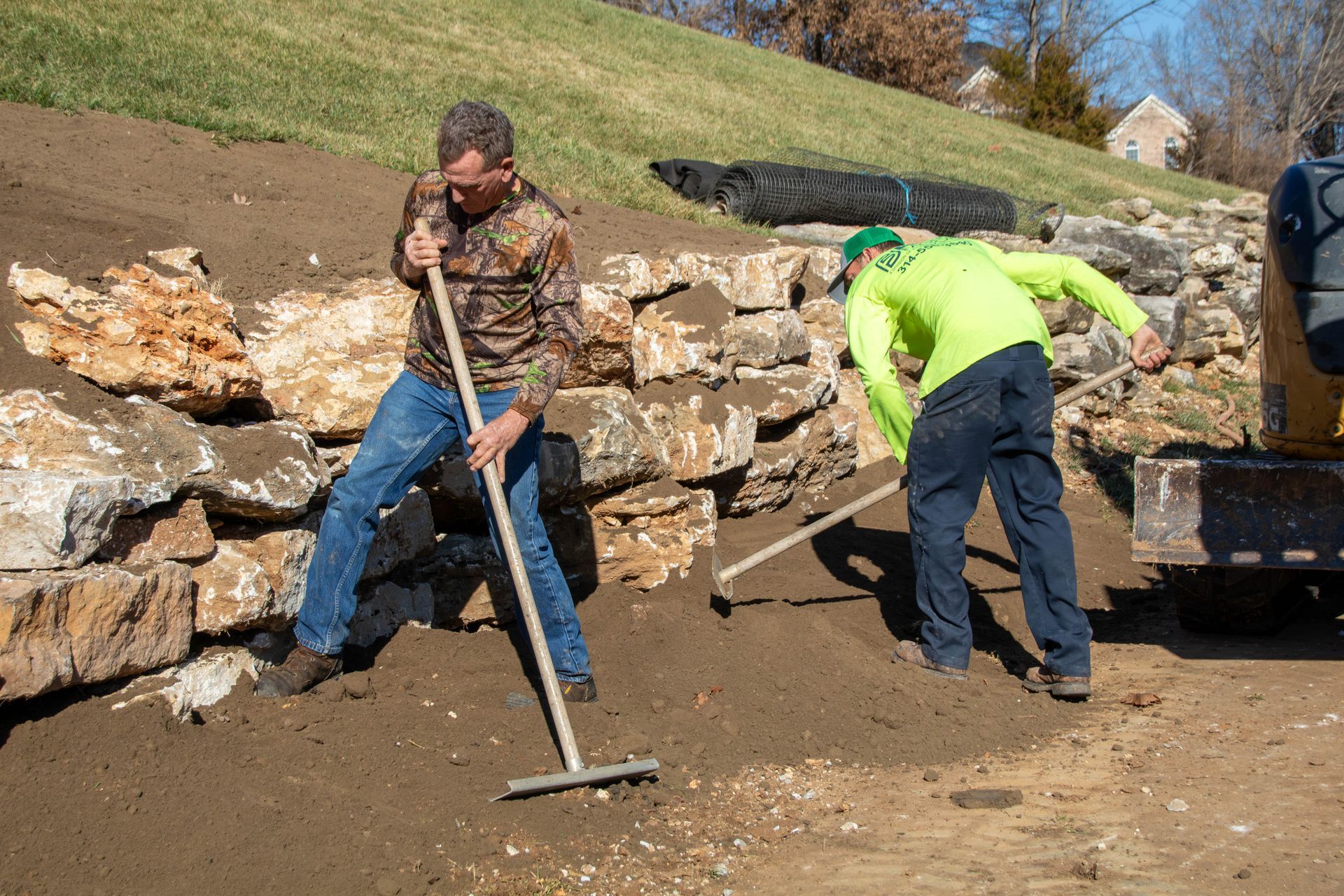 Two people raking soil near a stone retaining wall on a sunny day.