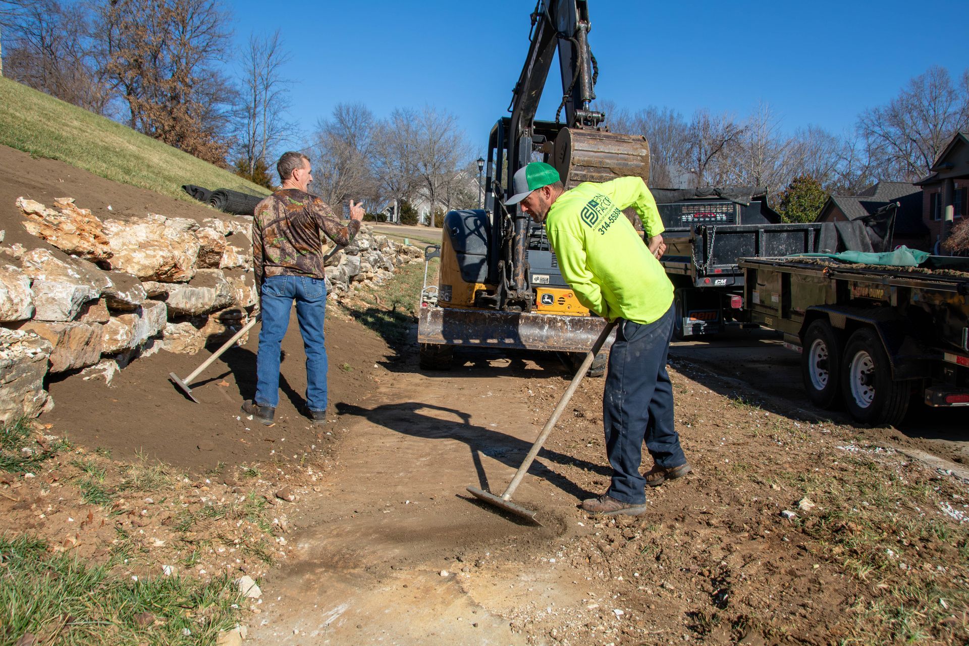 Two workers raking dirt on a construction site with an excavator and retaining wall.