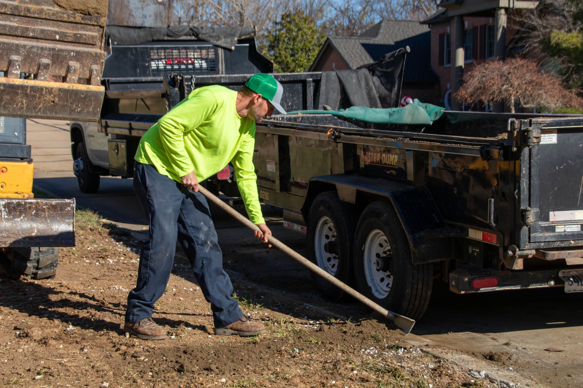 Man in safety vest shovels dirt near a trailer and equipment on a roadside.