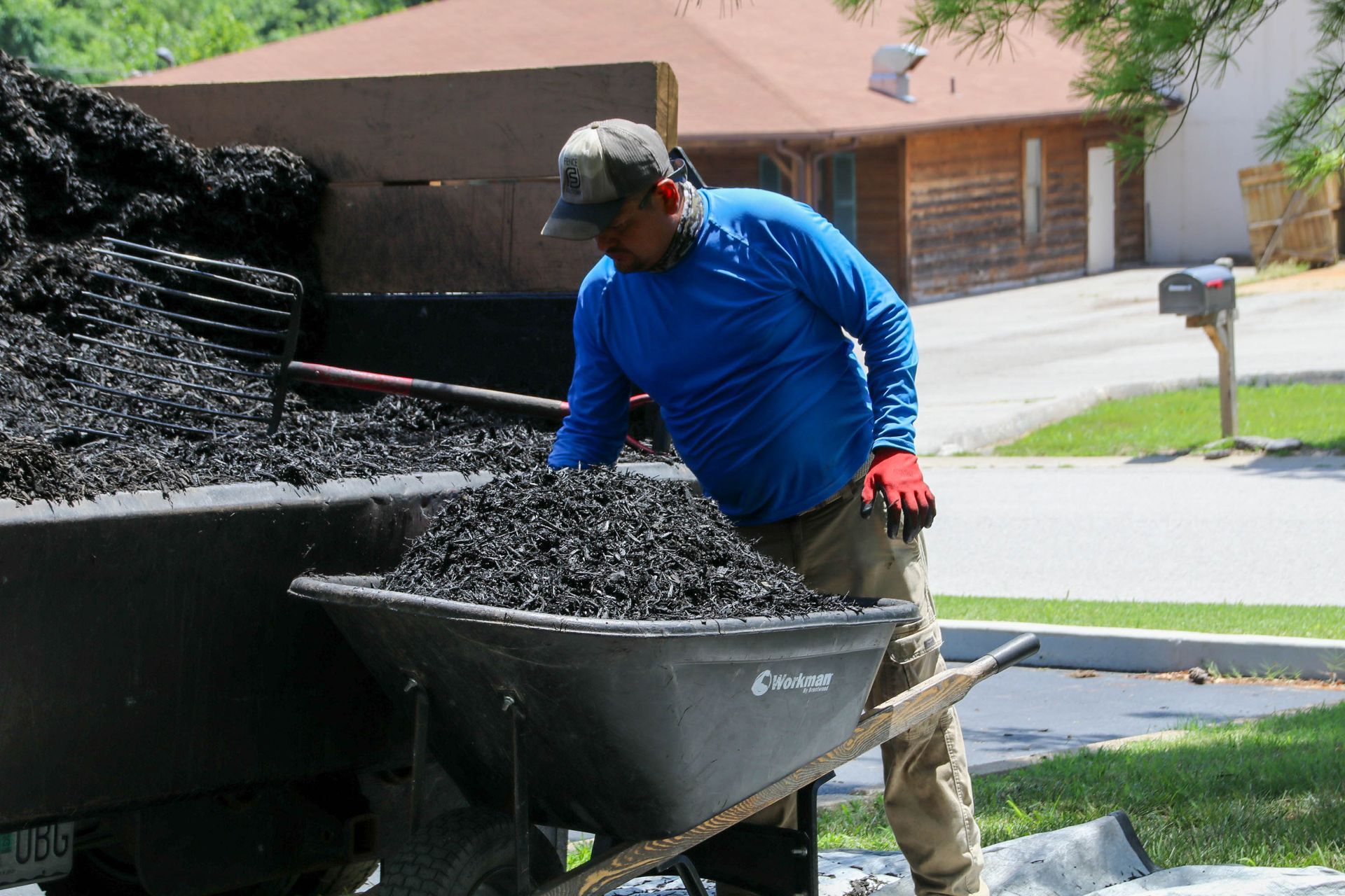 Man loading black asphalt into a wheelbarrow from a truck, outdoors on a sunny day.