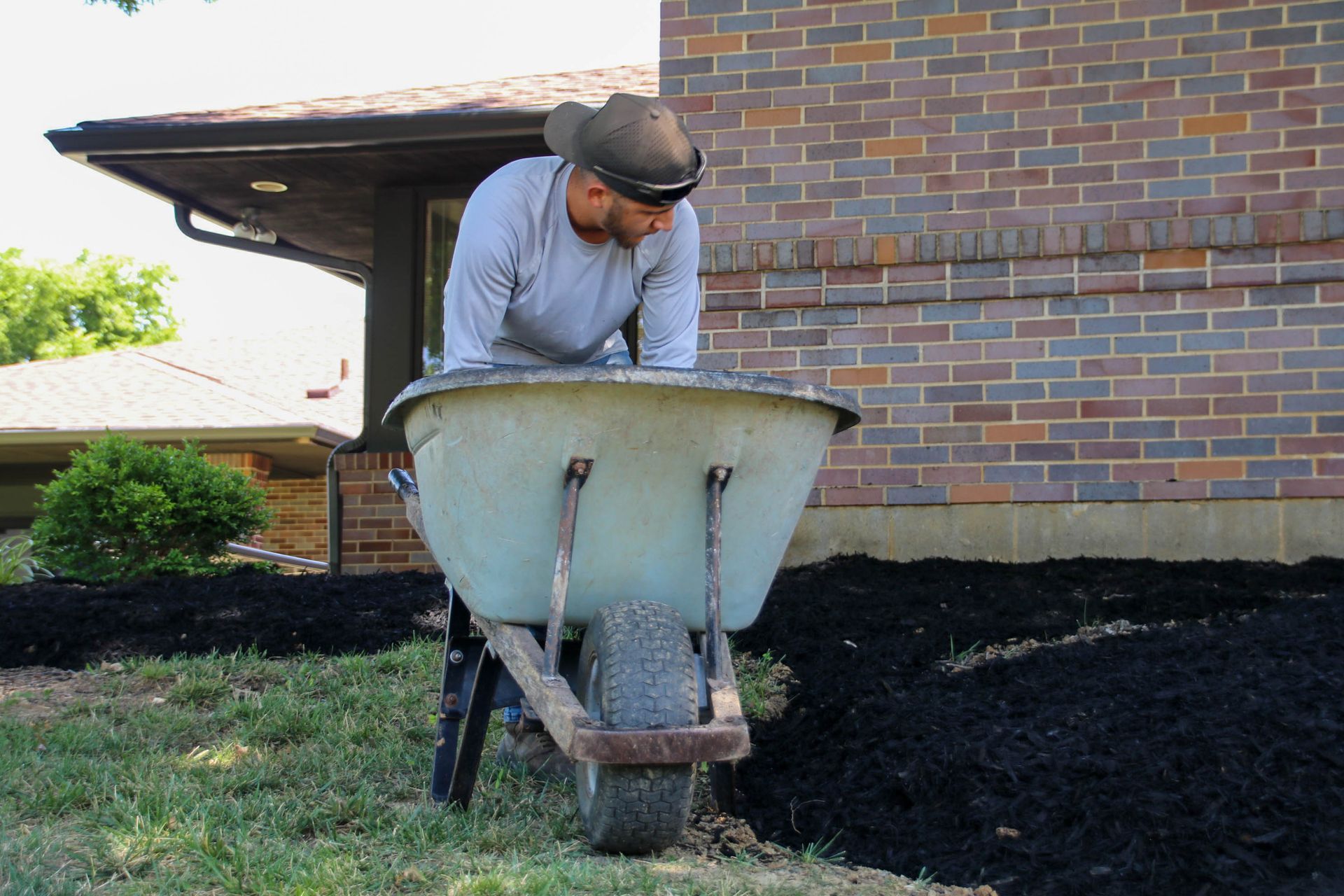 Man leaning over a wheelbarrow filled with dark mulch, near a brick house.