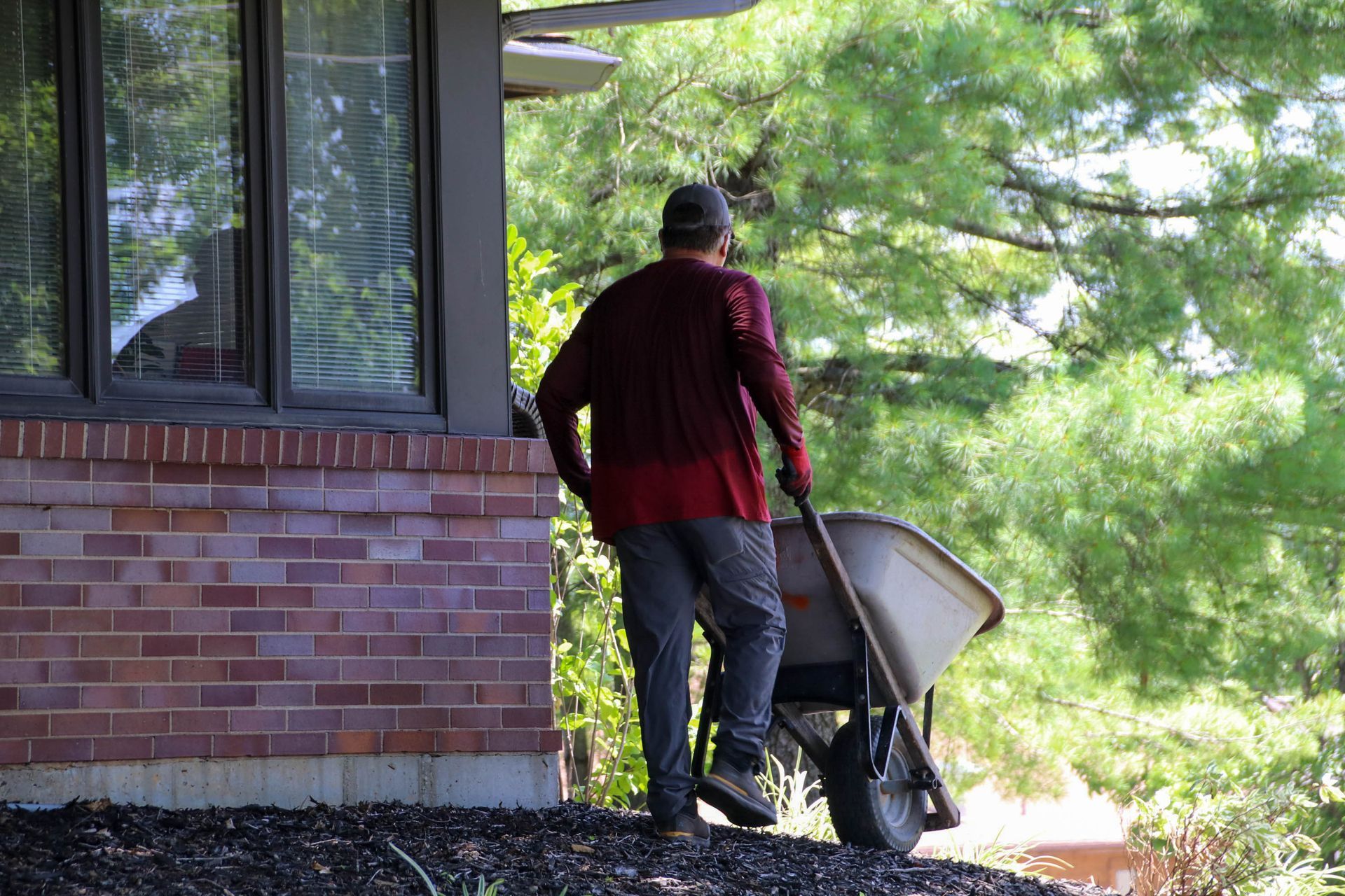 Person pushing a wheelbarrow near a brick building. Trees and windows are in the background.