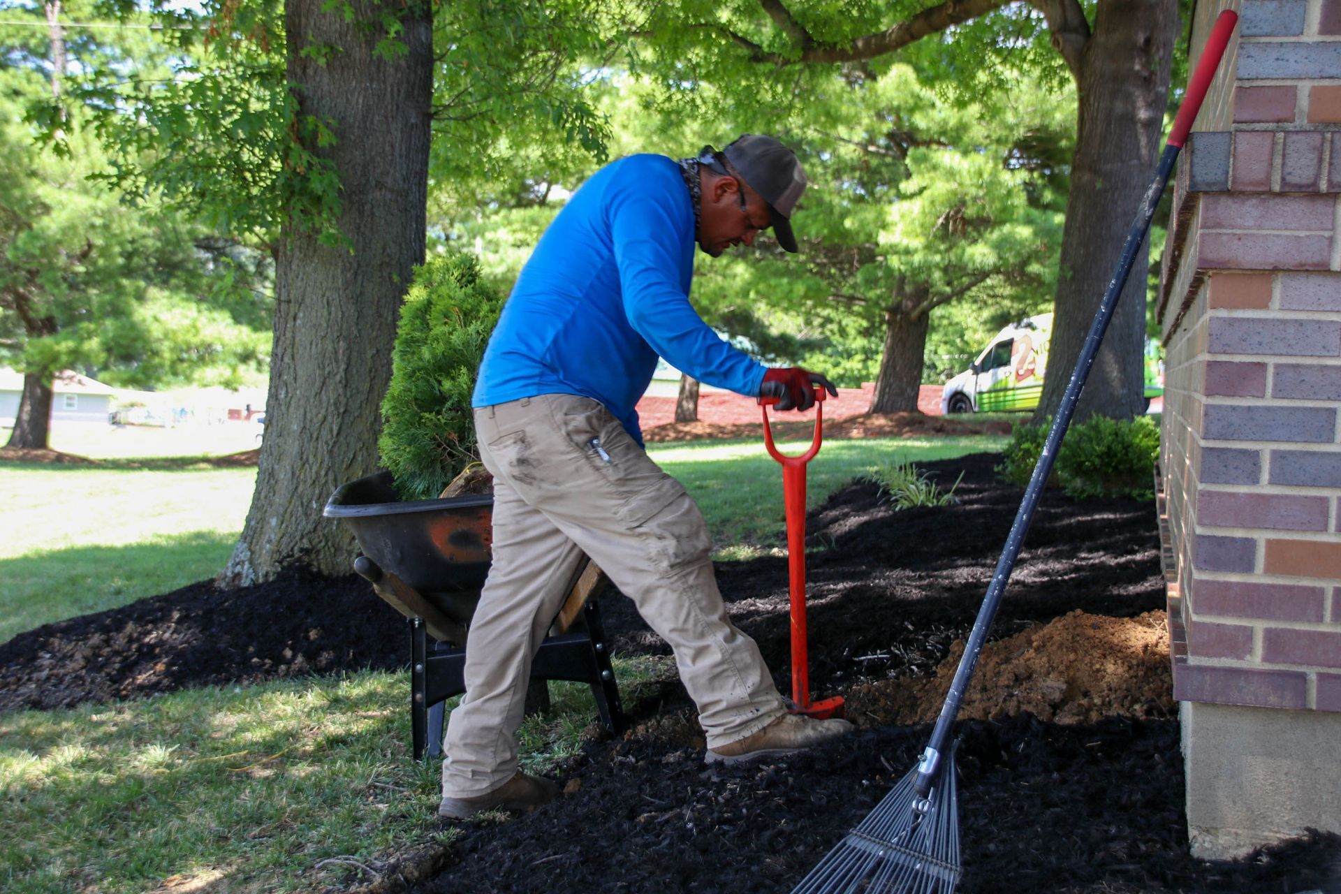 Man using a shovel in a mulch bed next to a building and a tree.
