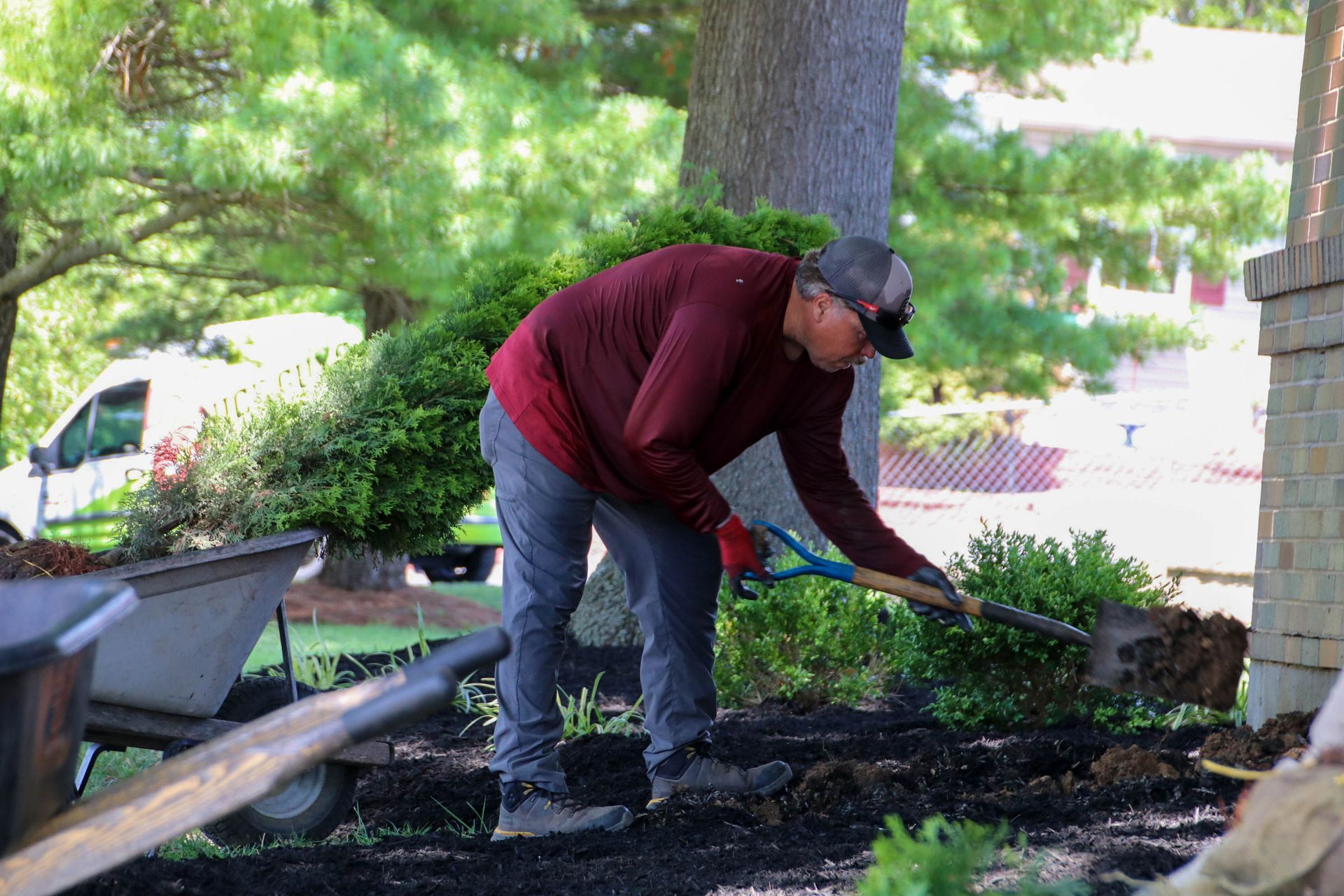 Man using a shovel to work in a garden near a building; mulch visible.