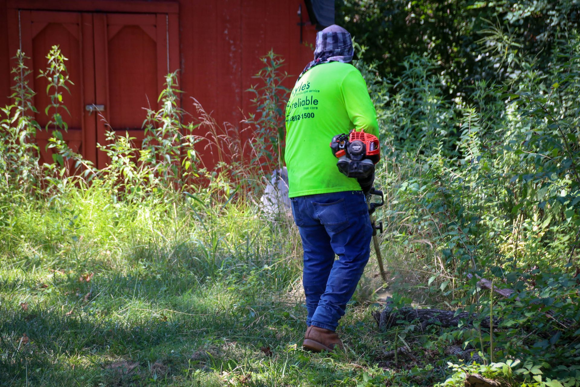 Person in neon green shirt using a weed whacker near a red building and overgrown vegetation.