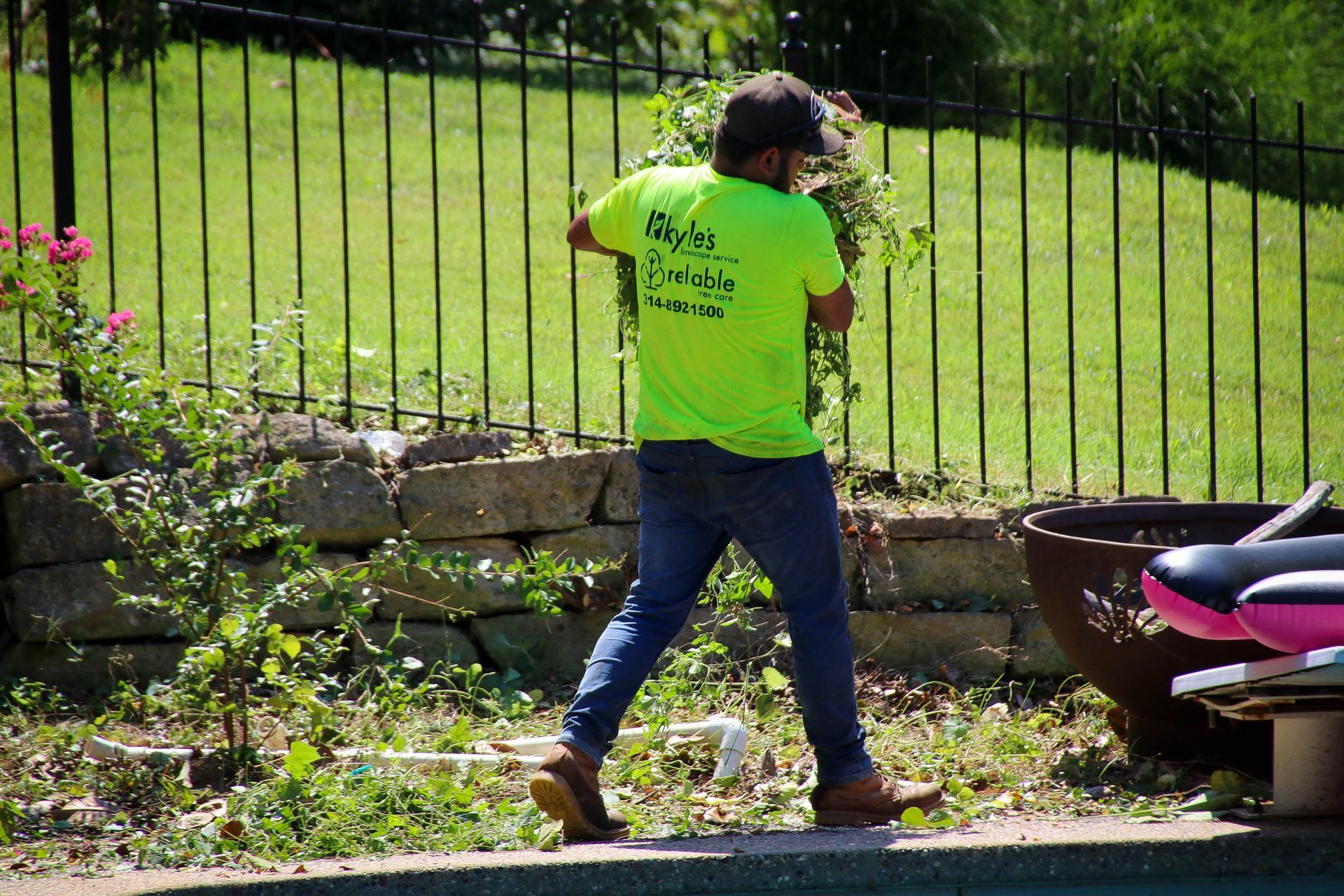 Man in neon shirt trimming bushes near a pool and black fence on a sunny day.
