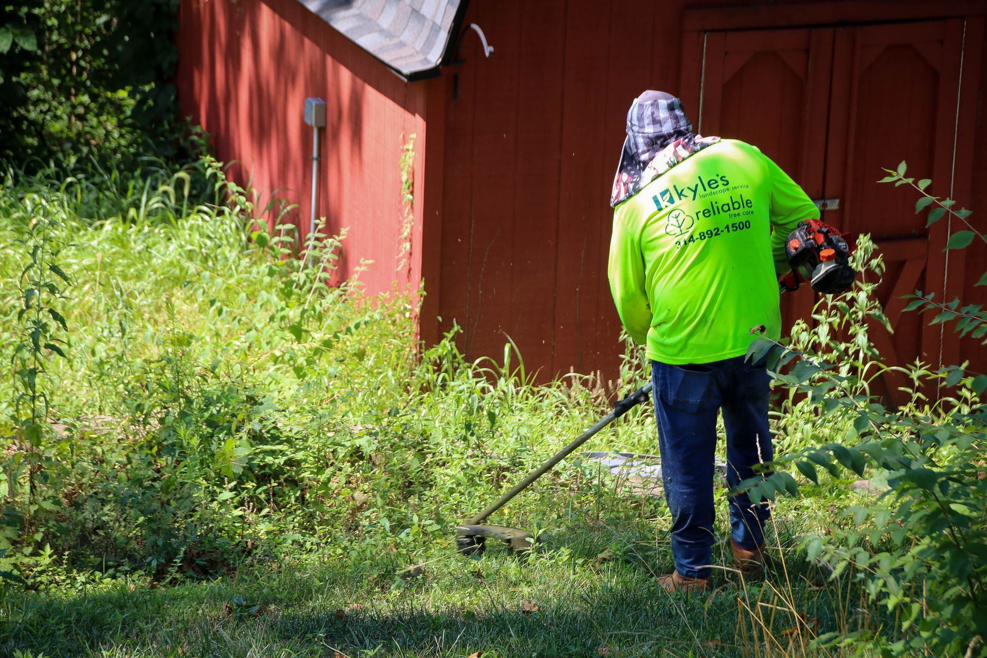 Person in neon shirt using a weed whacker near a red shed, cutting overgrown weeds and grass.