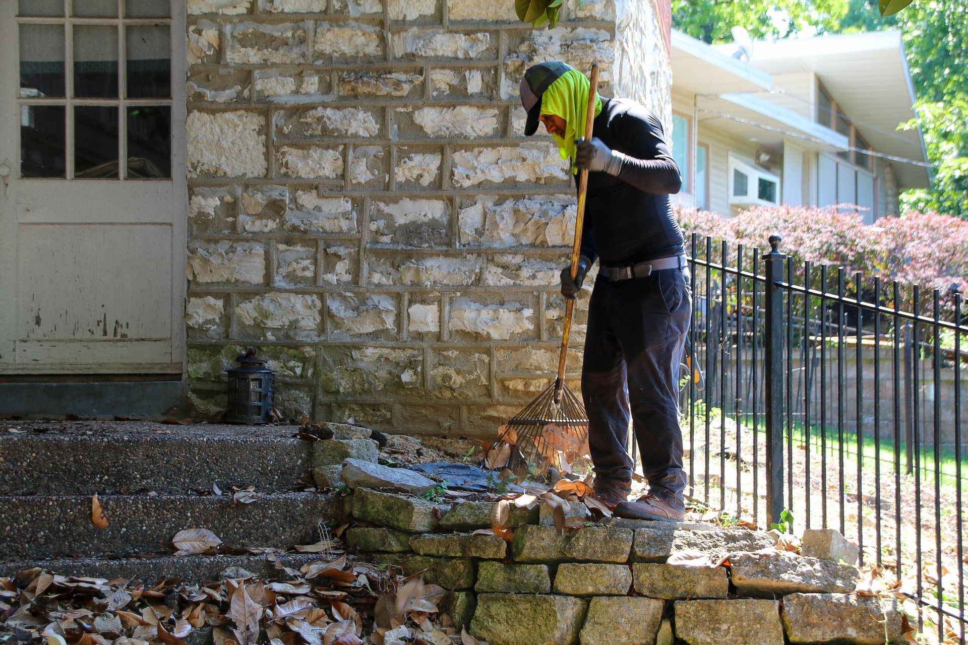 Person in safety vest rakes debris from a stone wall near a building.
