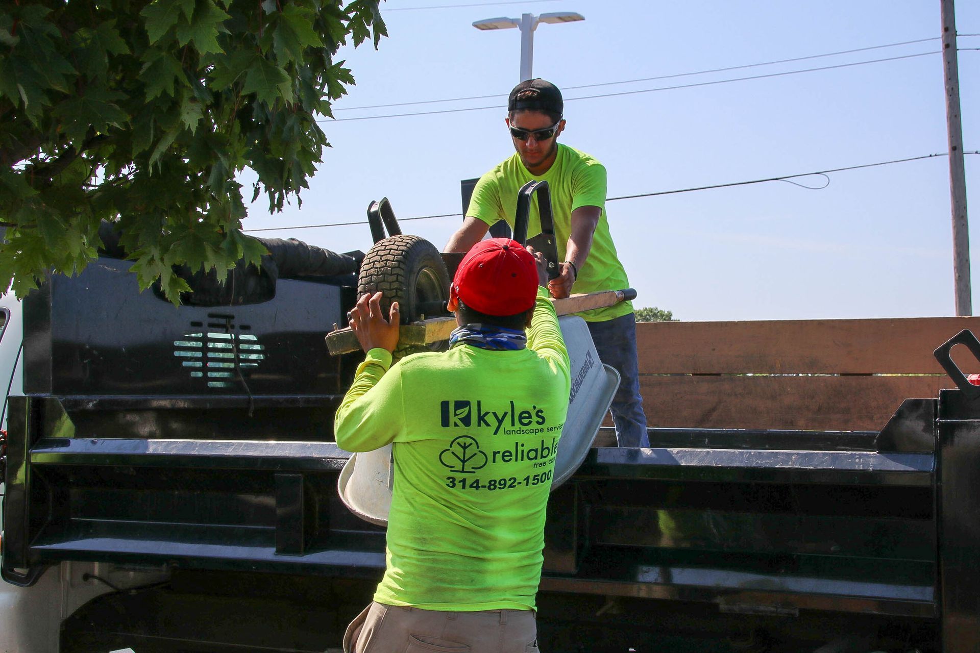 Two workers in green shirts loading equipment into the back of a black truck on a sunny day.