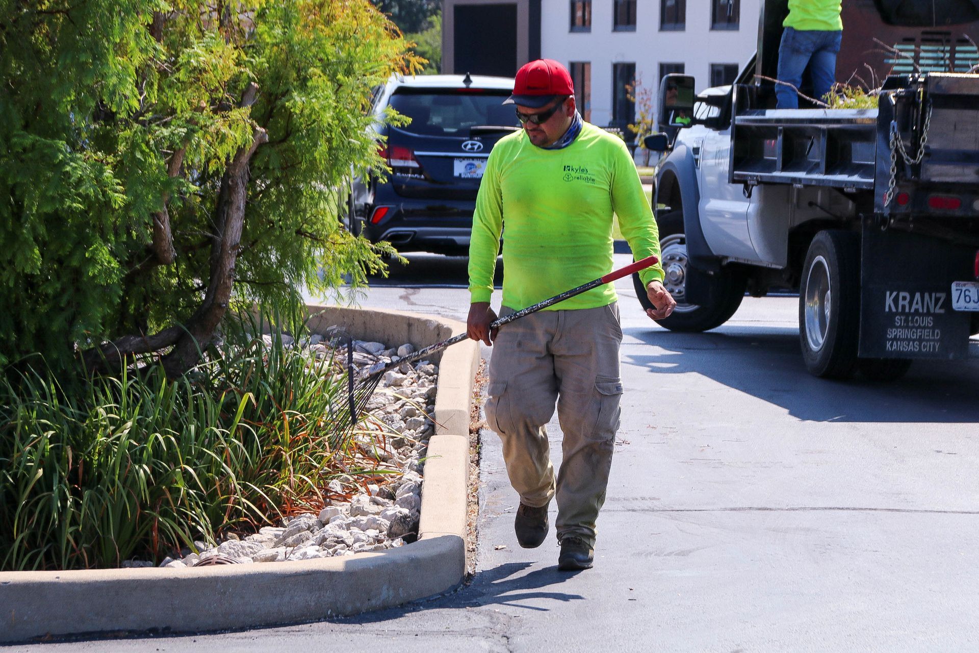 Man in neon green shirt walking next to curb, holding a rod. Landscaping truck in background.