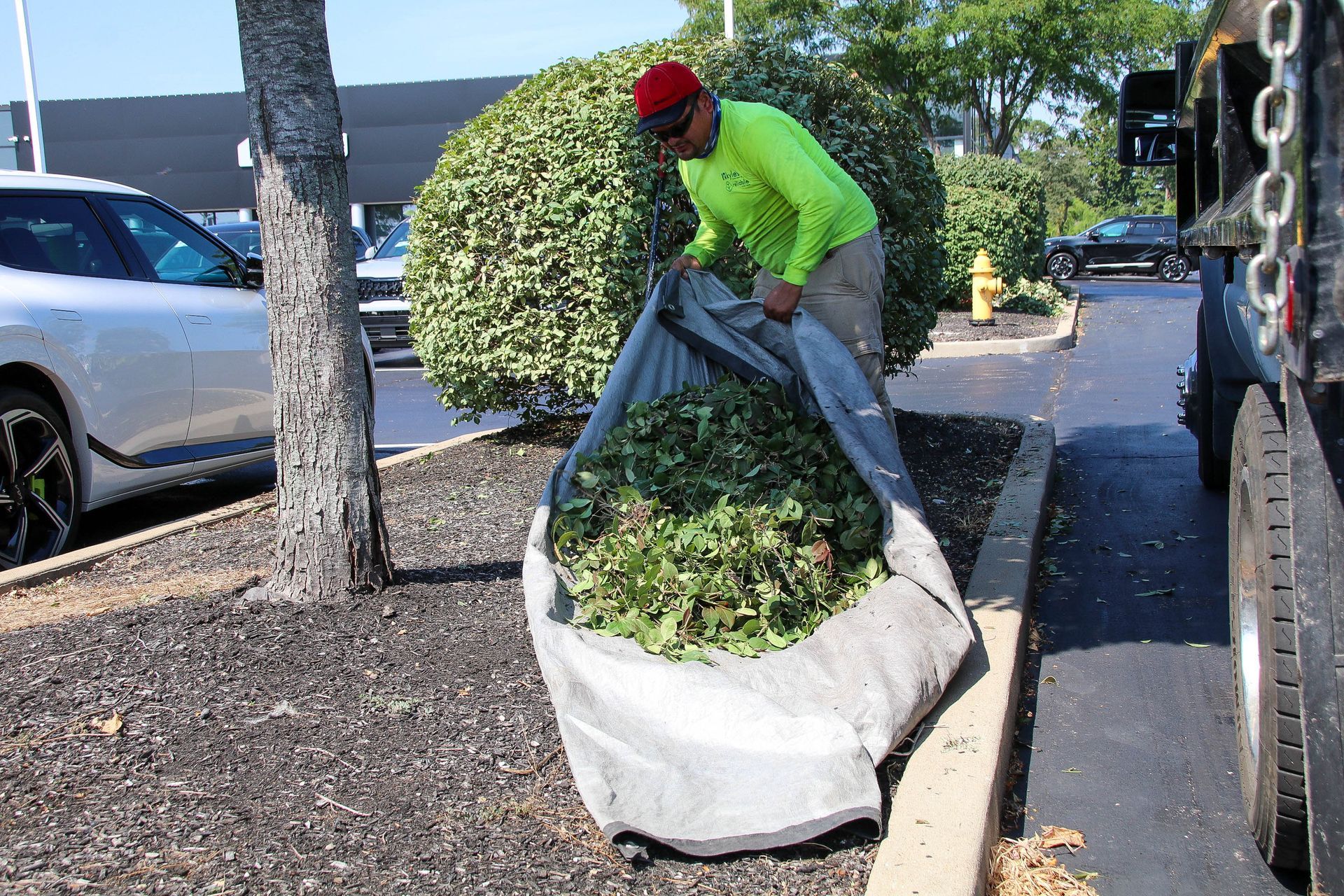 Man in neon shirt filling a large bag with yard waste next to a truck in a parking lot.