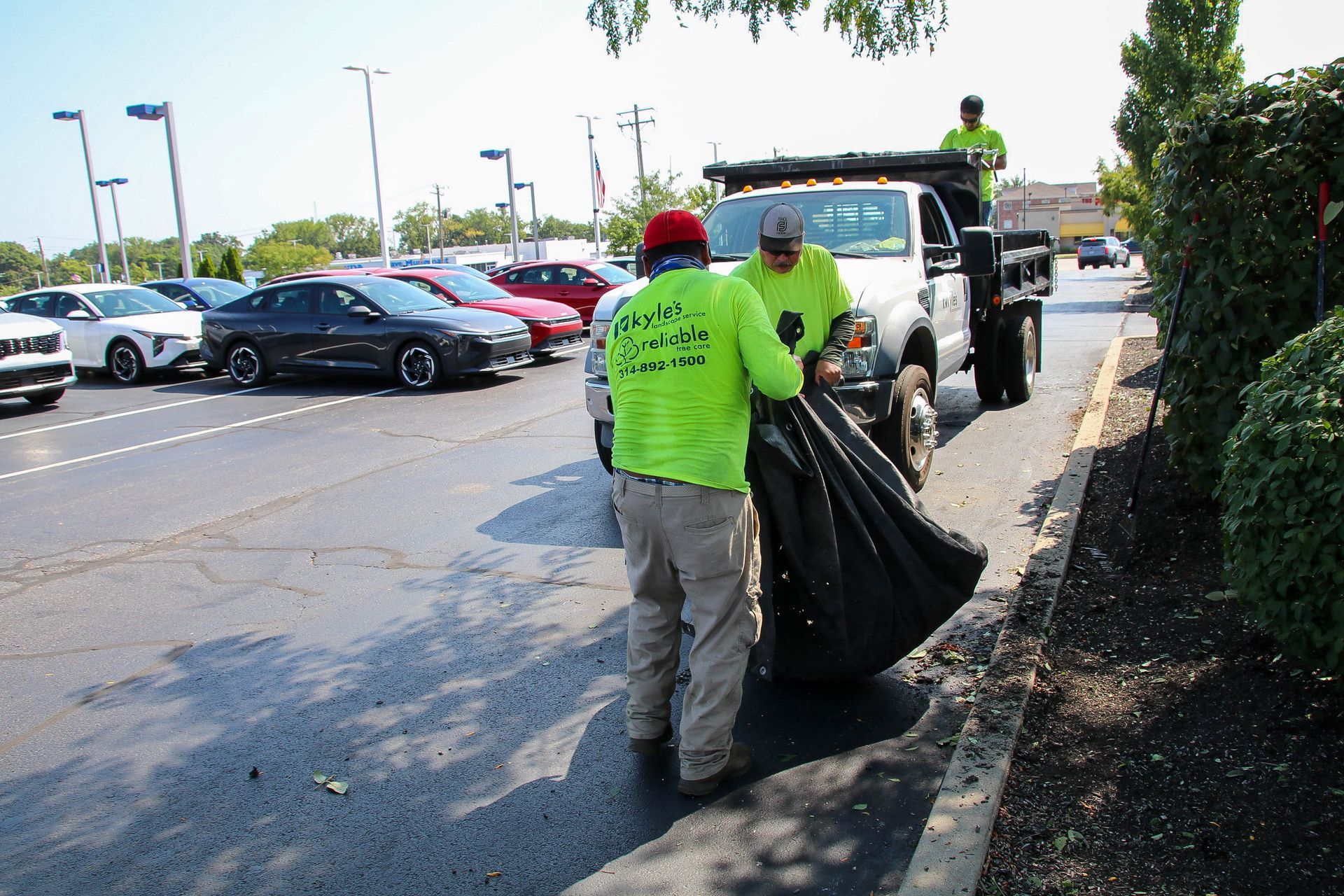 Two people in neon green shirts collecting trash into a black bag, near a parked truck and cars.
