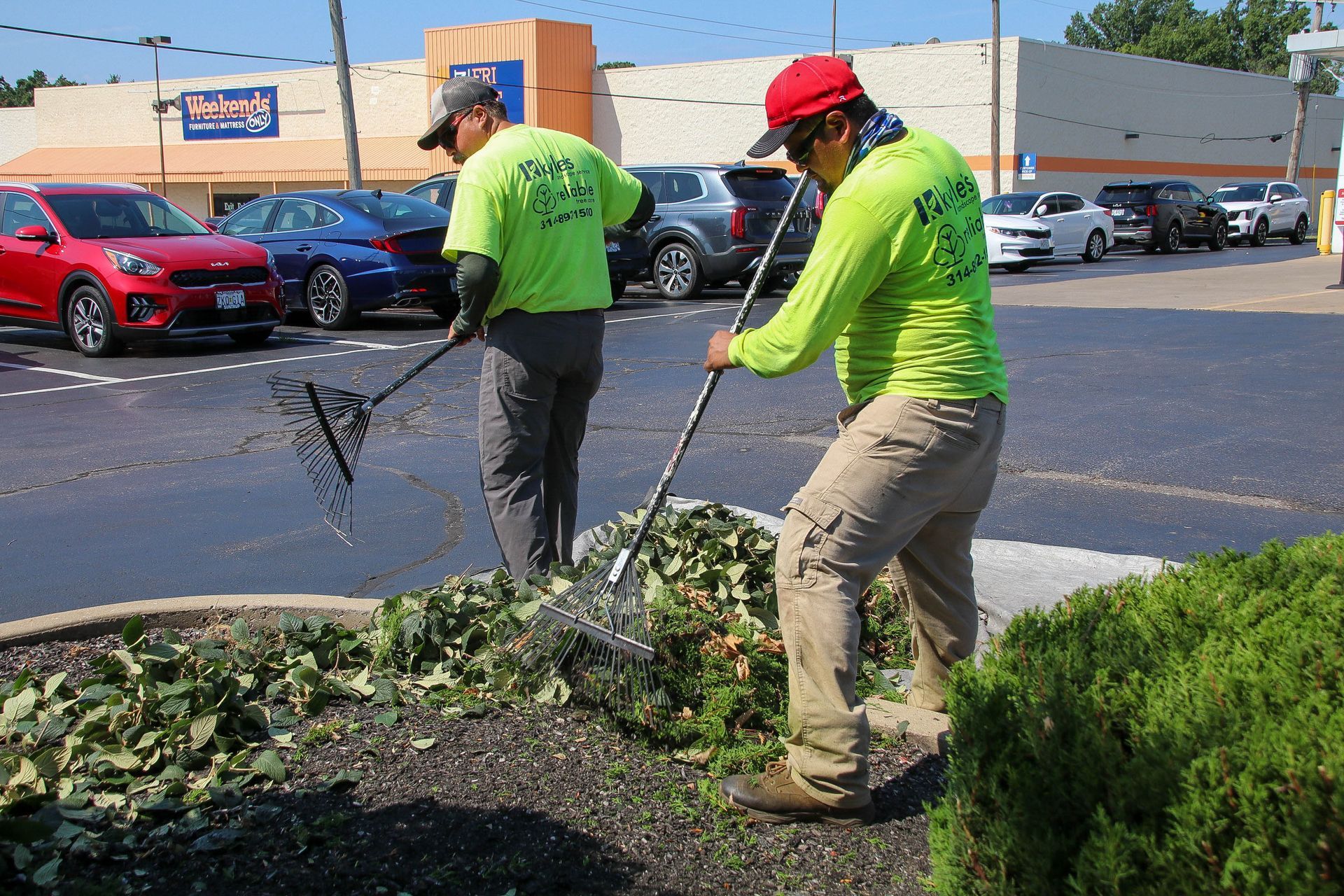 Two workers in neon green shirts raking leaves in a landscaped area near a parking lot.