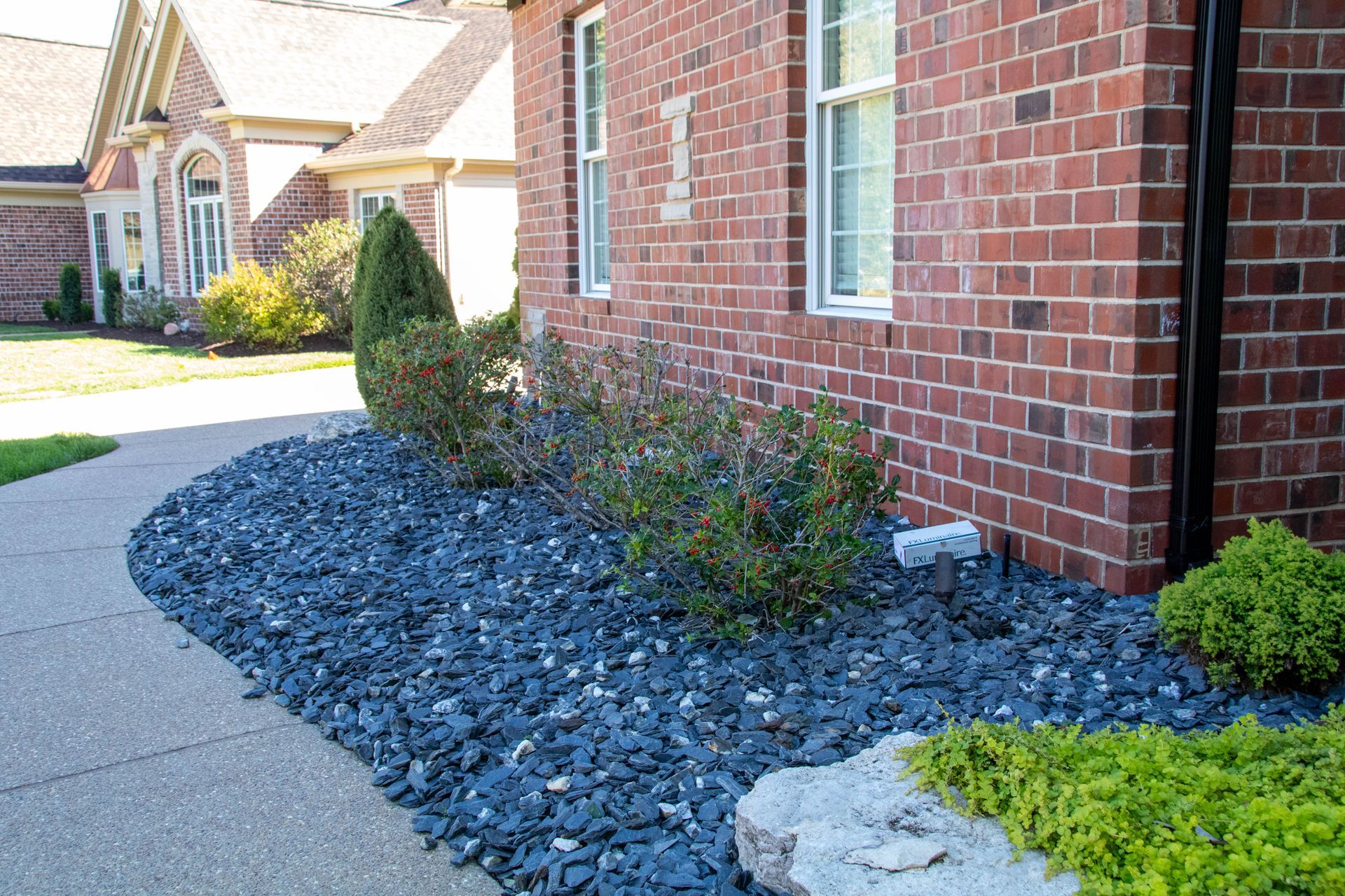 Sidewalk curves past a brick house, a bed of dark blue rocks, and shrubs.