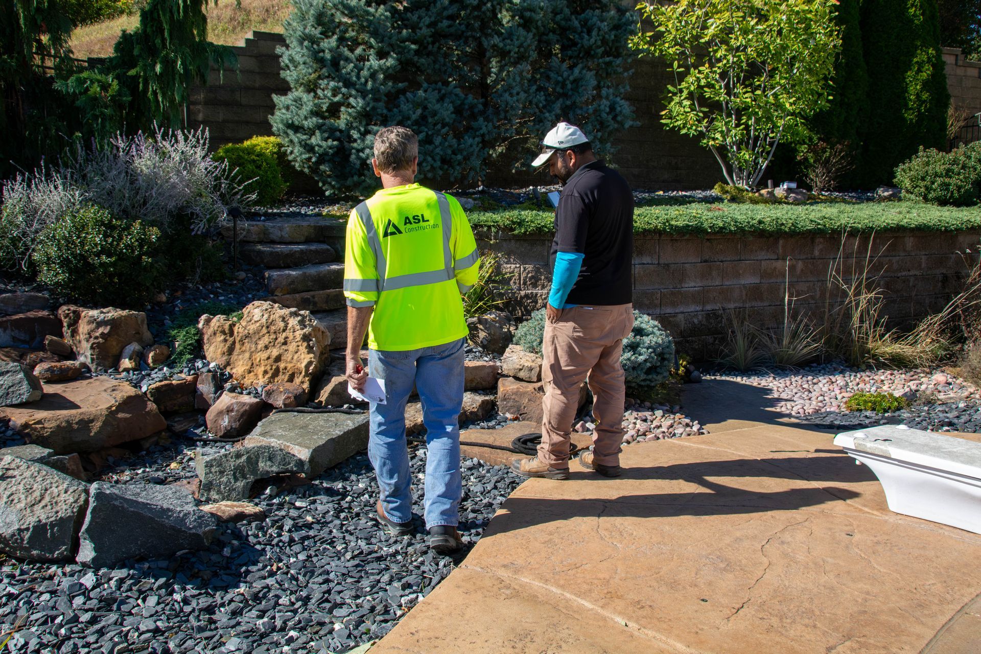Two people, one in a neon vest, looking at a landscaped area with rocks and a concrete path.