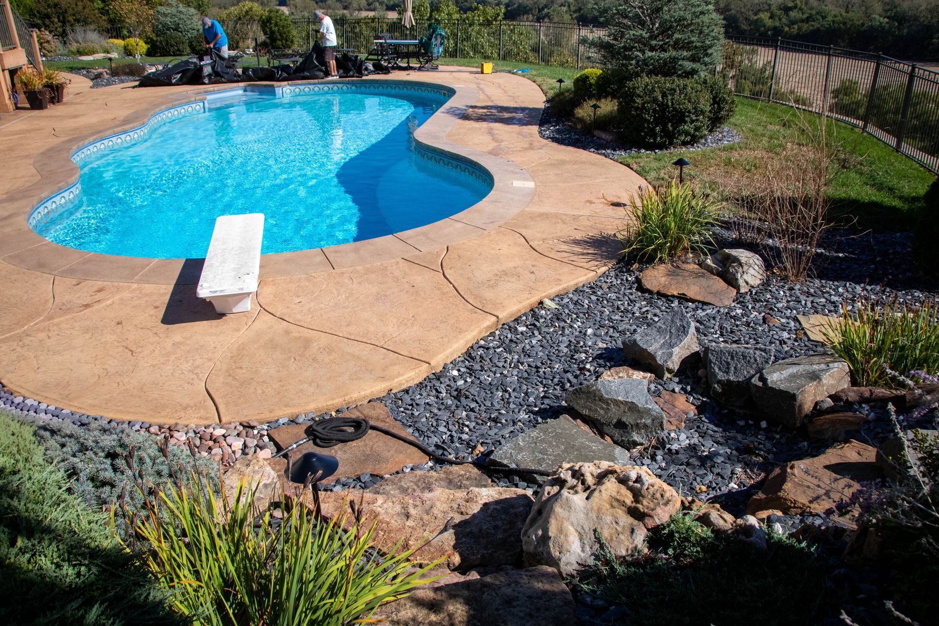 Pool with diving board and decorative rock garden. Landscaped backyard with people.