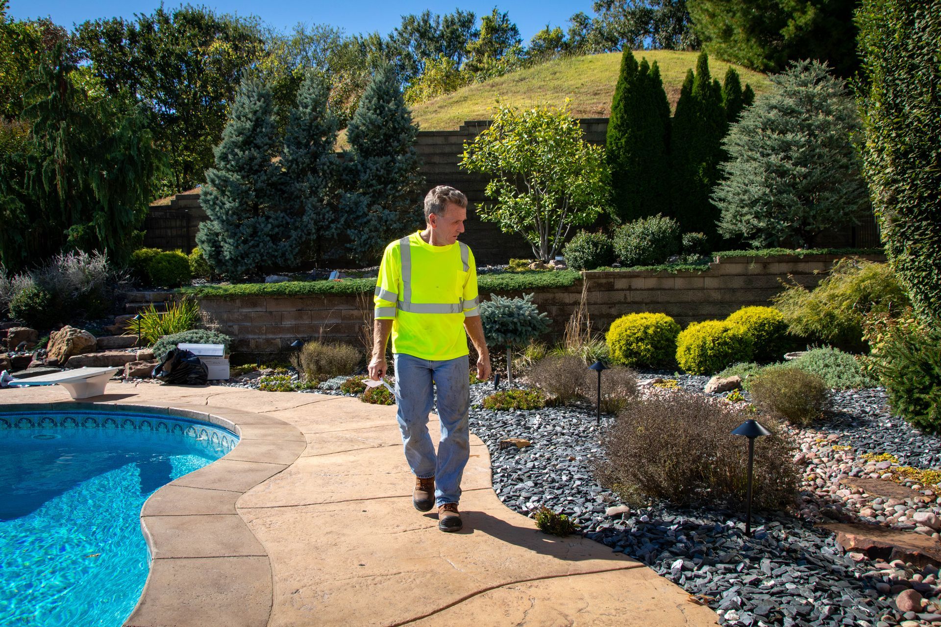 Man in reflective vest walks along a pool path in a landscaped backyard.