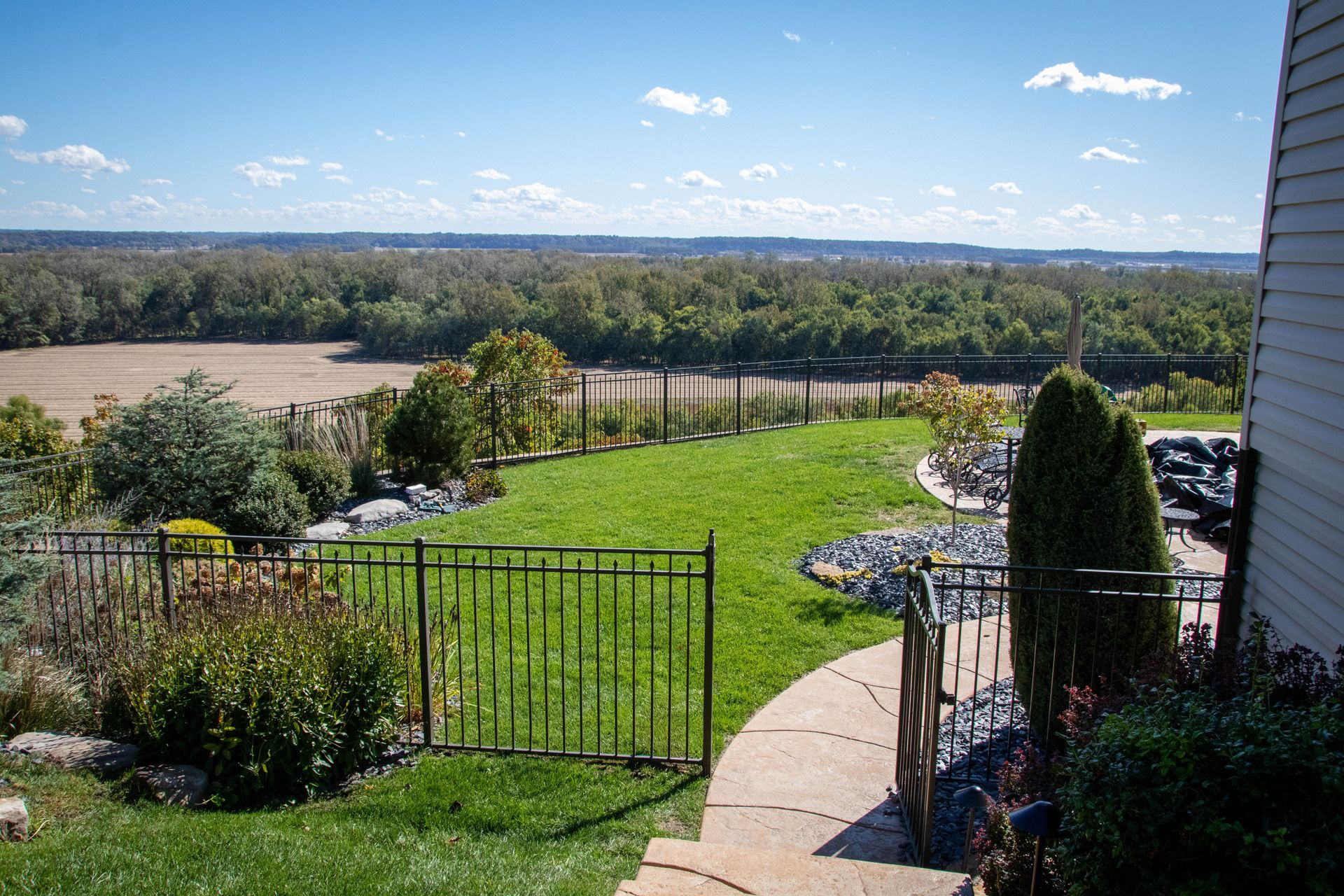 View from a house's yard: green lawn, black fence, trees in the distance, blue sky.