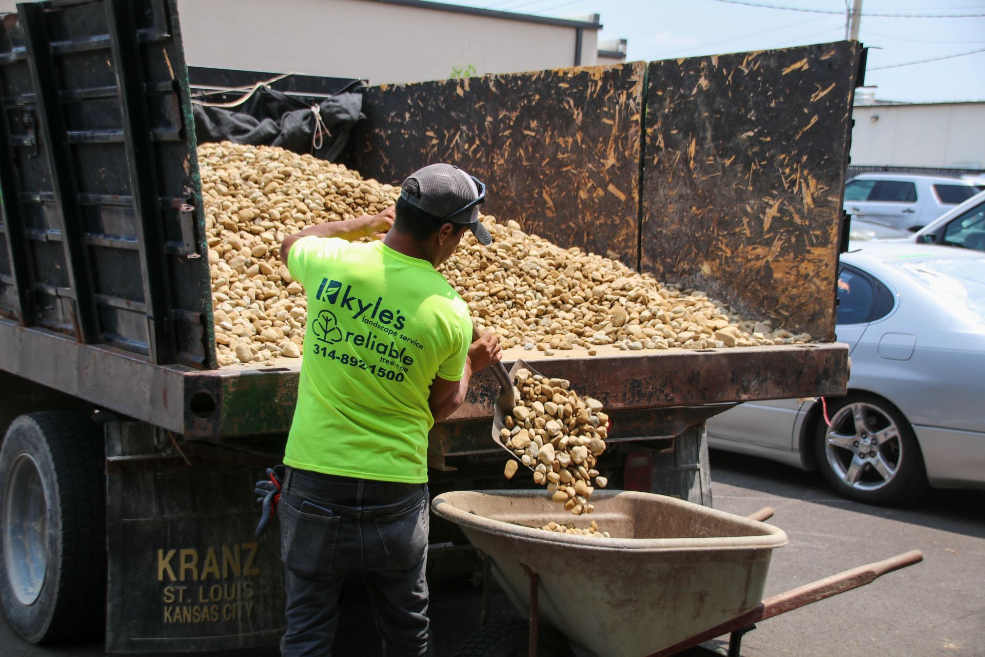 Man in green shirt shoveling rocks from a dump truck into a wheelbarrow.