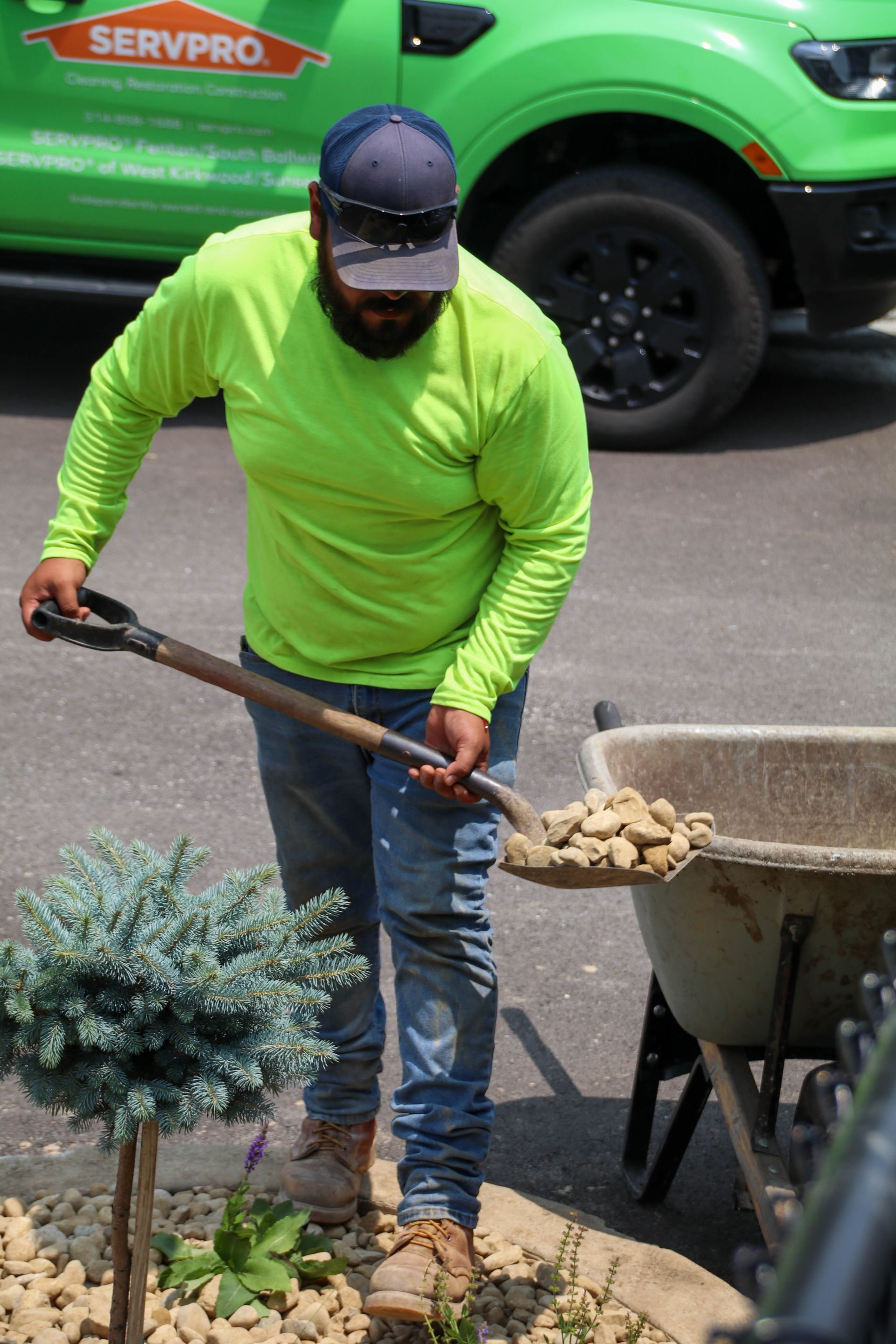 Man in neon green shirt shovels rocks into wheelbarrow. Servpro truck in background.