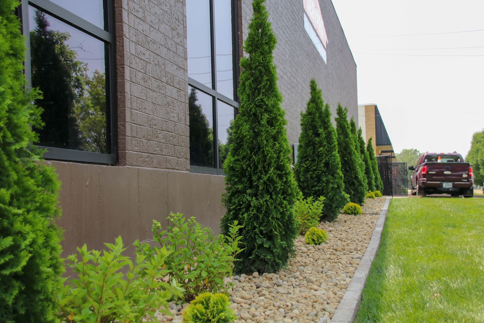 Green trees and bushes border a brown brick building; a red pickup truck is visible.