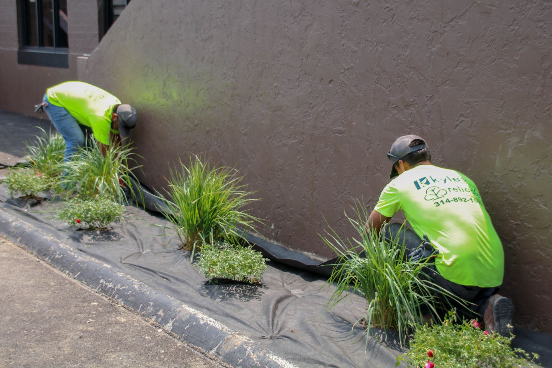 Two people in neon shirts planting in a garden bed next to a brown wall.