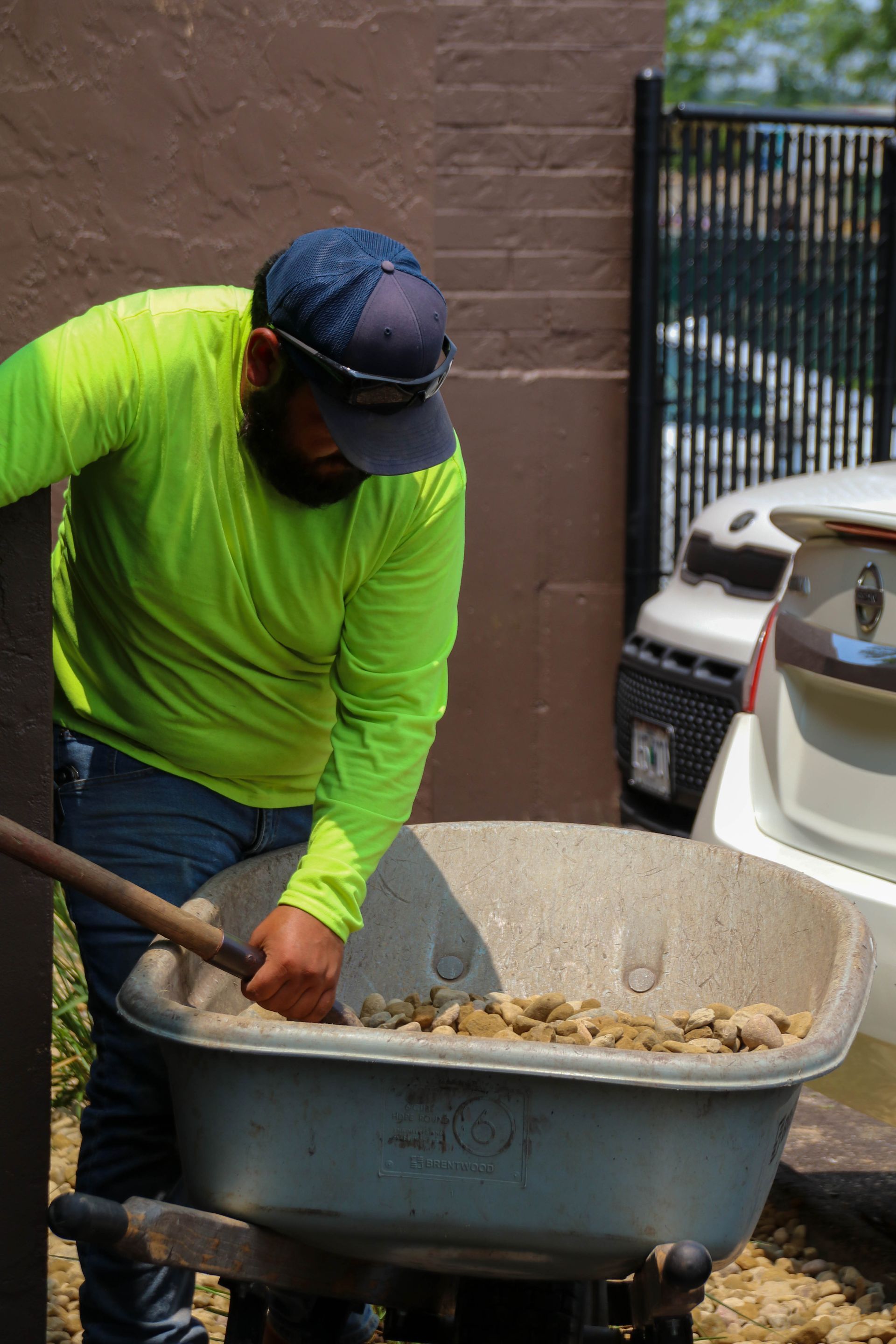 A person in a neon green shirt uses a shovel to move rocks in a wheelbarrow outdoors.