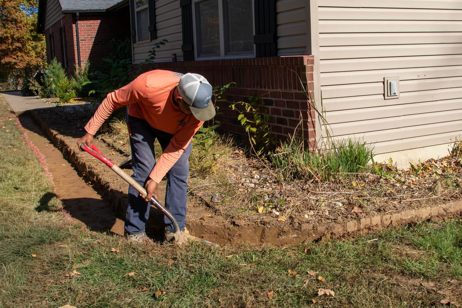 Person digging a trench in front of a house. He wears an orange shirt and a hat.
