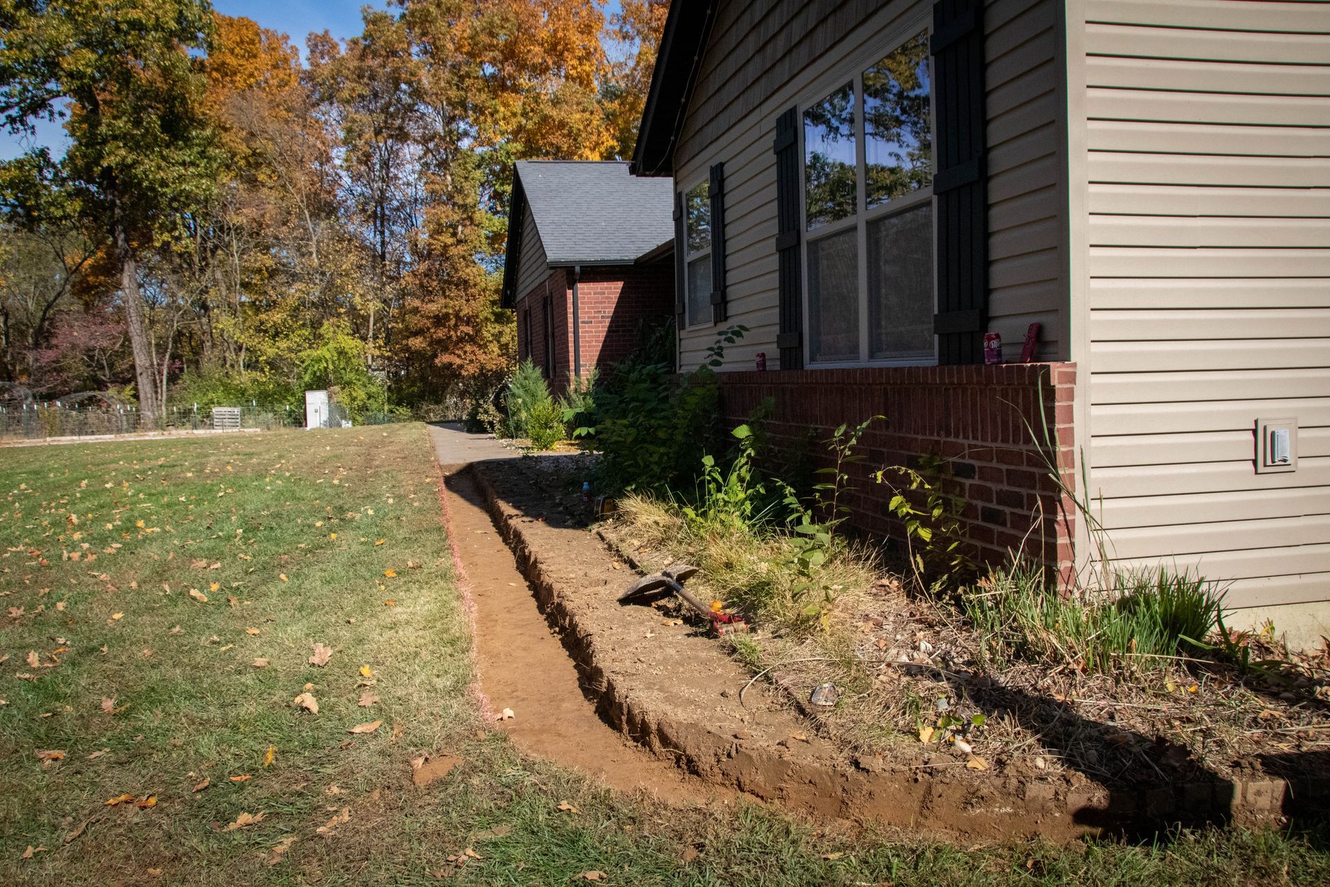 Trench dug along a house foundation in a yard with fall foliage in the background.