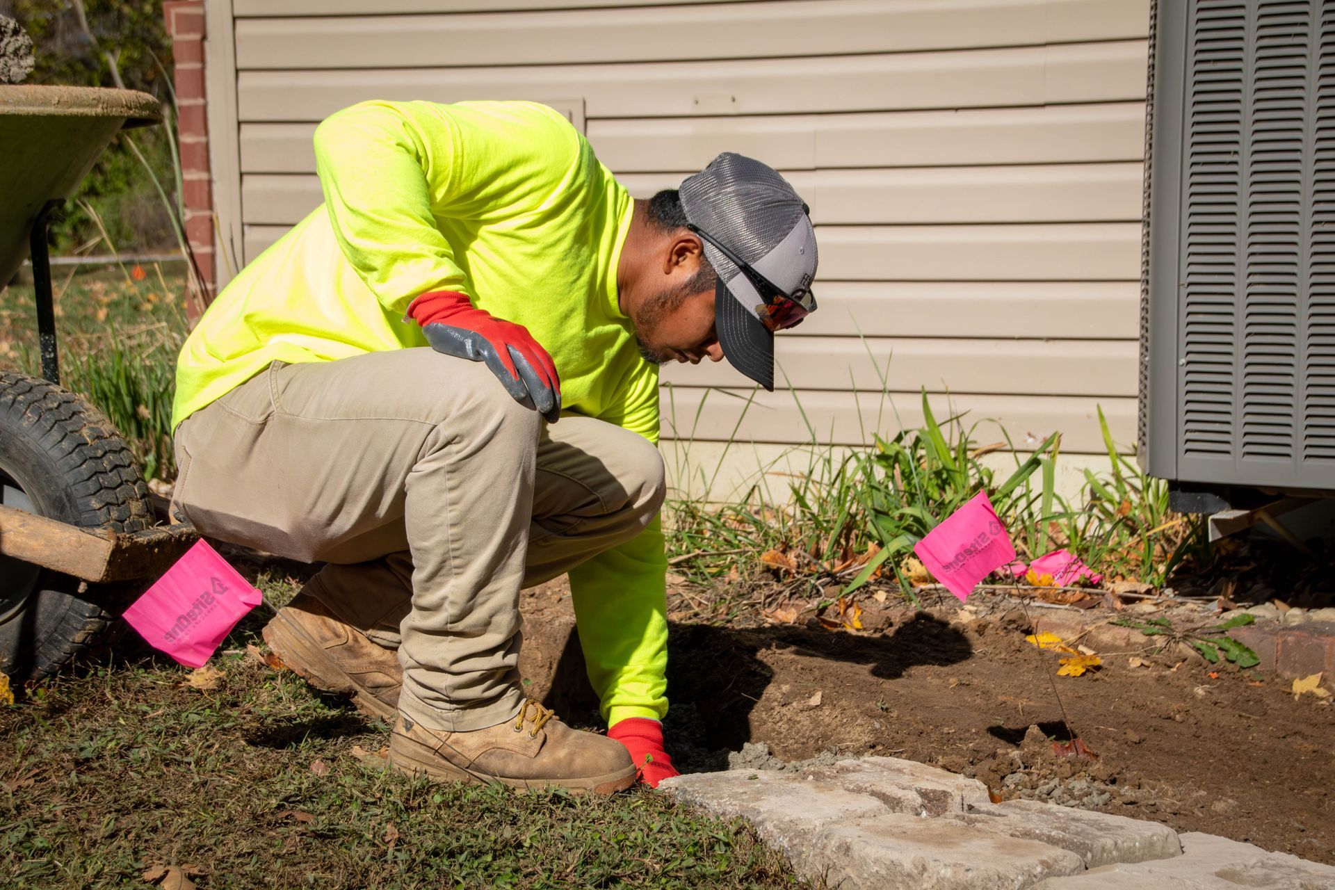 Landscaper in neon yellow shirt, kneeling, placing paving stones near a building, next to a wheelbarrow and markers.