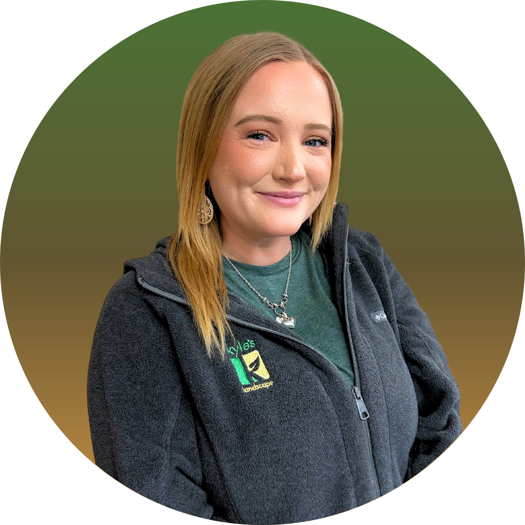 Woman with blonde hair wearing a green shirt and dark gray zip-up. She smiles, with a green and brown background.