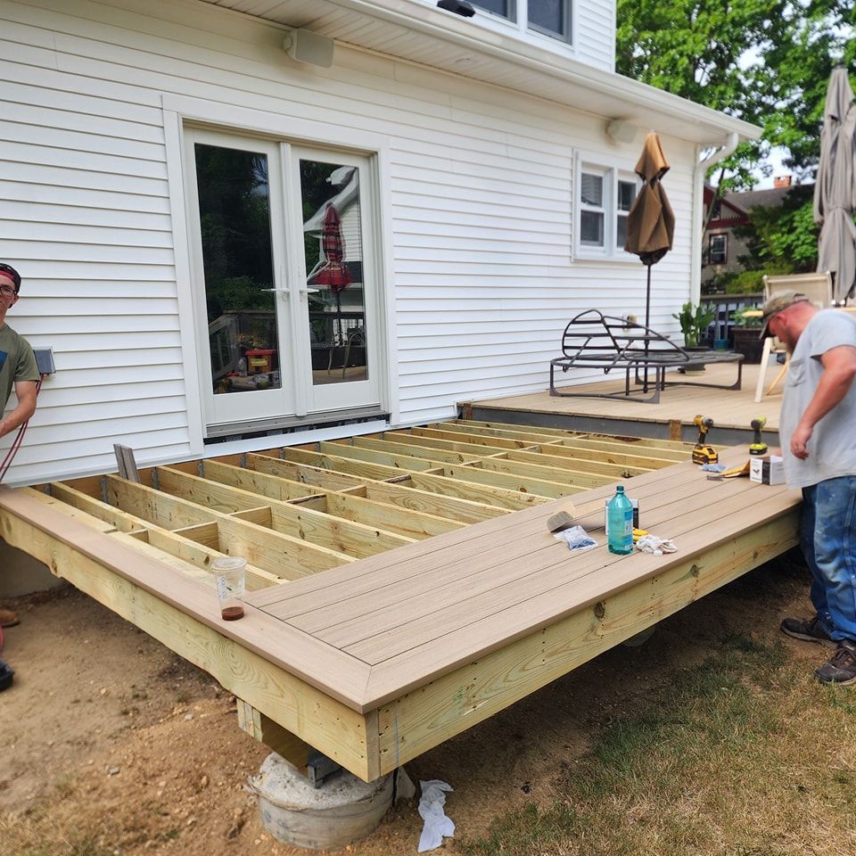Two men are working on a wooden deck in front of a white house