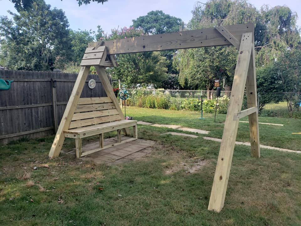 A wooden swing with a bench attached to it is sitting in the grass in a backyard.