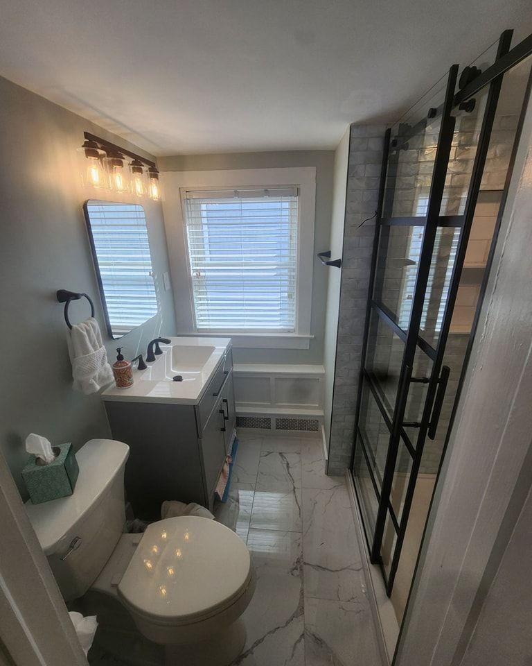 A bathroom with a toilet , sink , mirror and window.