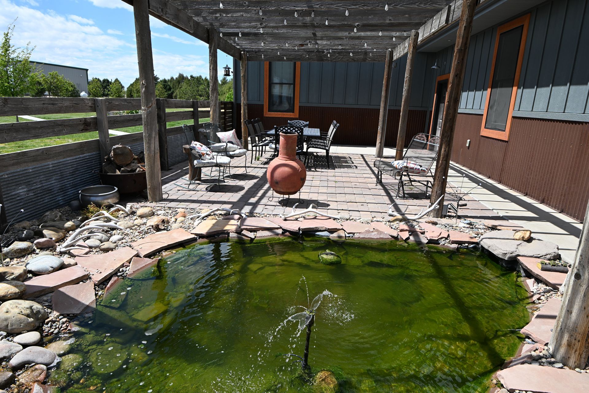 Green algae-filled backyard pool beside a patio with broken tiles and outdoor furniture under a pergola