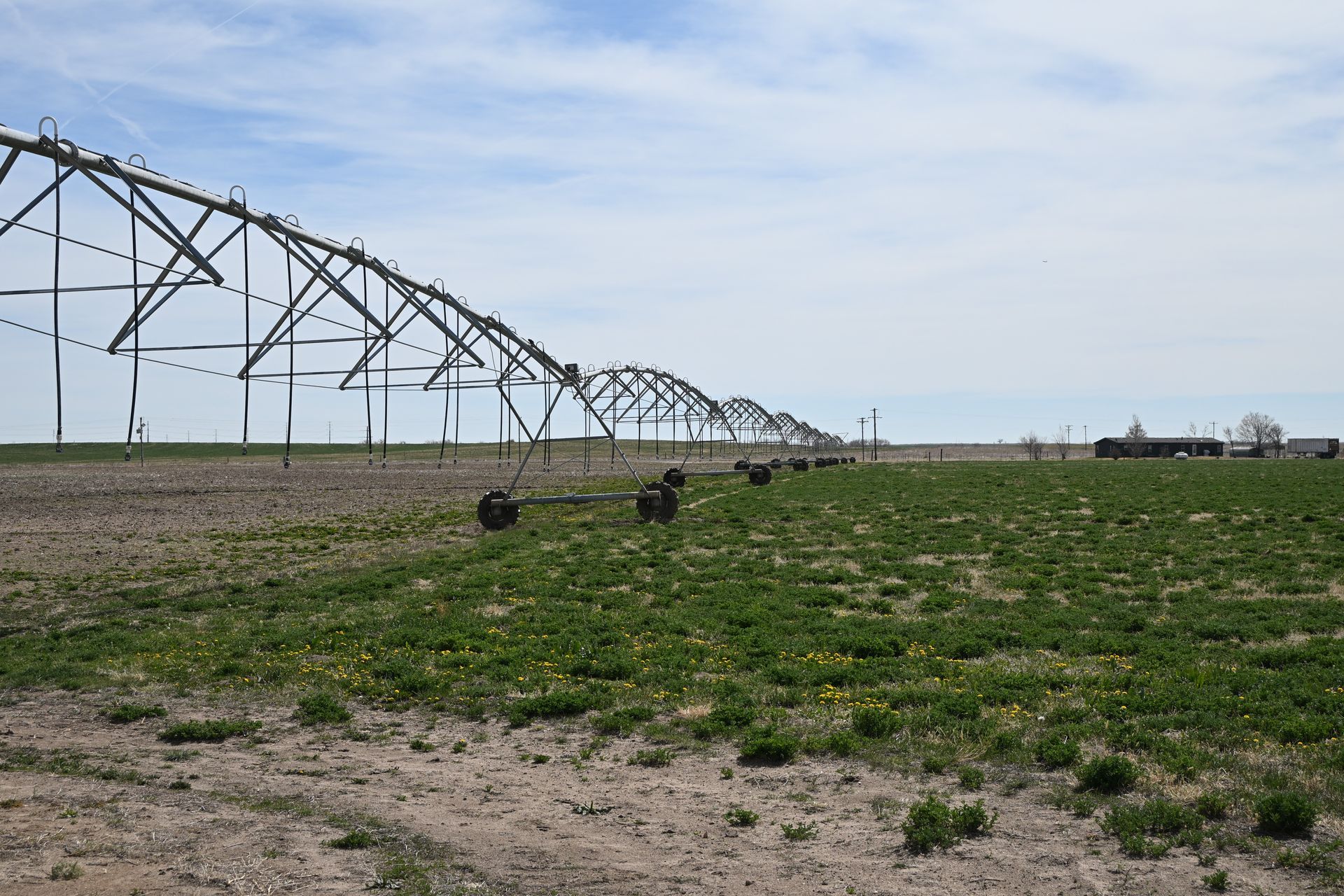Irrigation sprinklers watering a grassy field beside a long metal pivot system under a cloudy sky