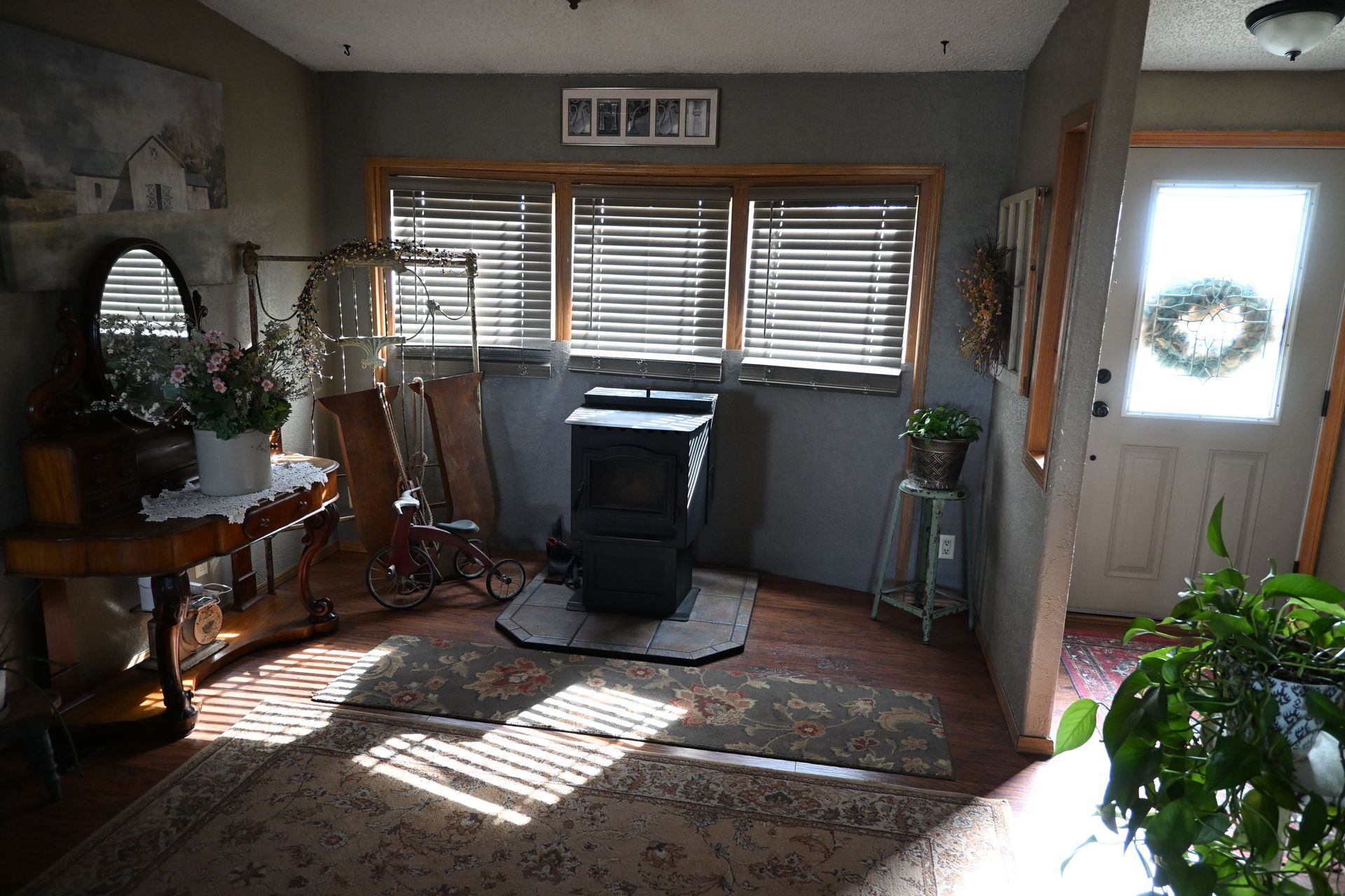 Sunlit living room with rug, potted plants, window blinds, and a front door wreath