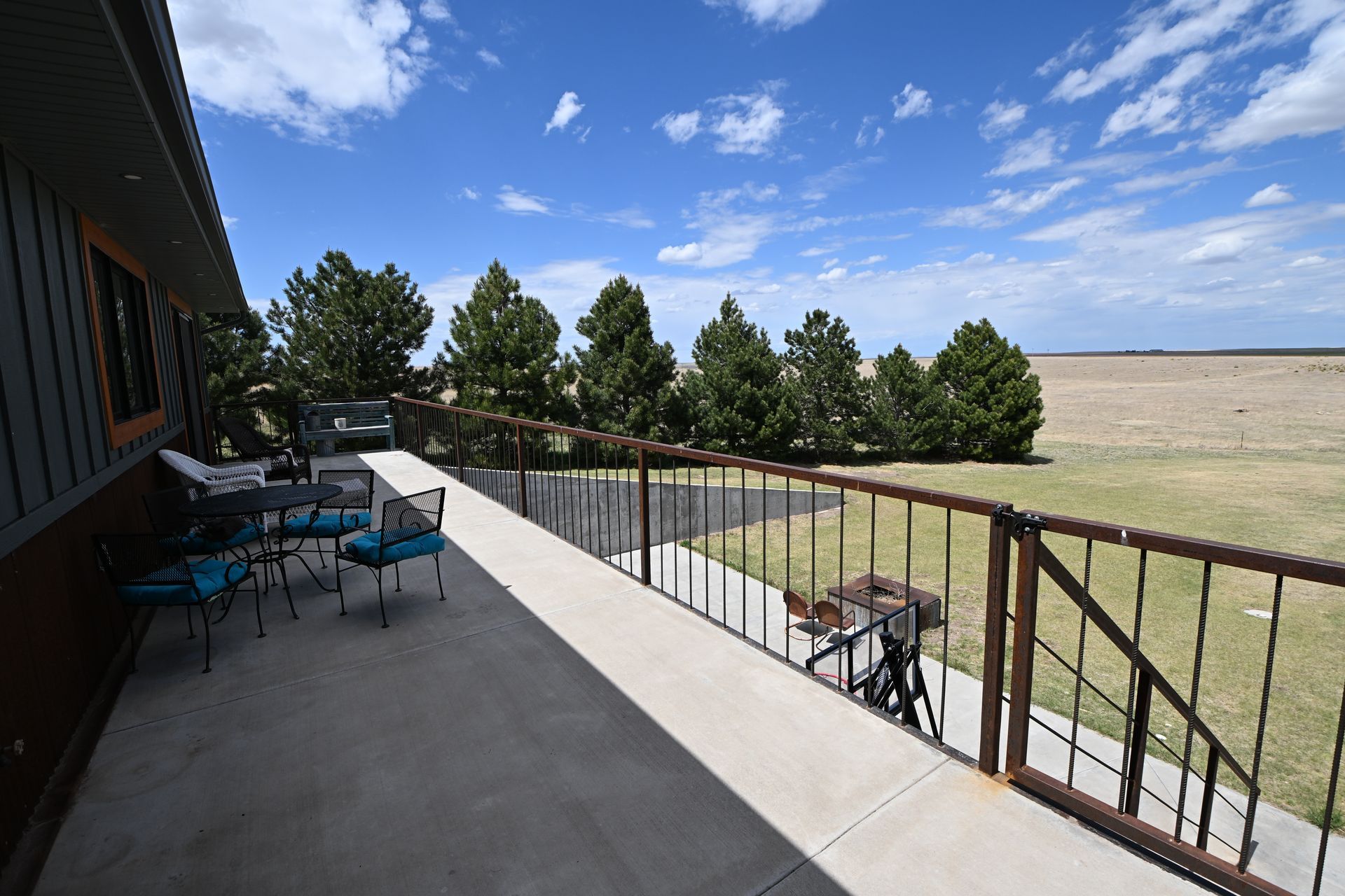 Patio with blue chairs and table beside a railing, overlooking an open field under a partly cloudy sky