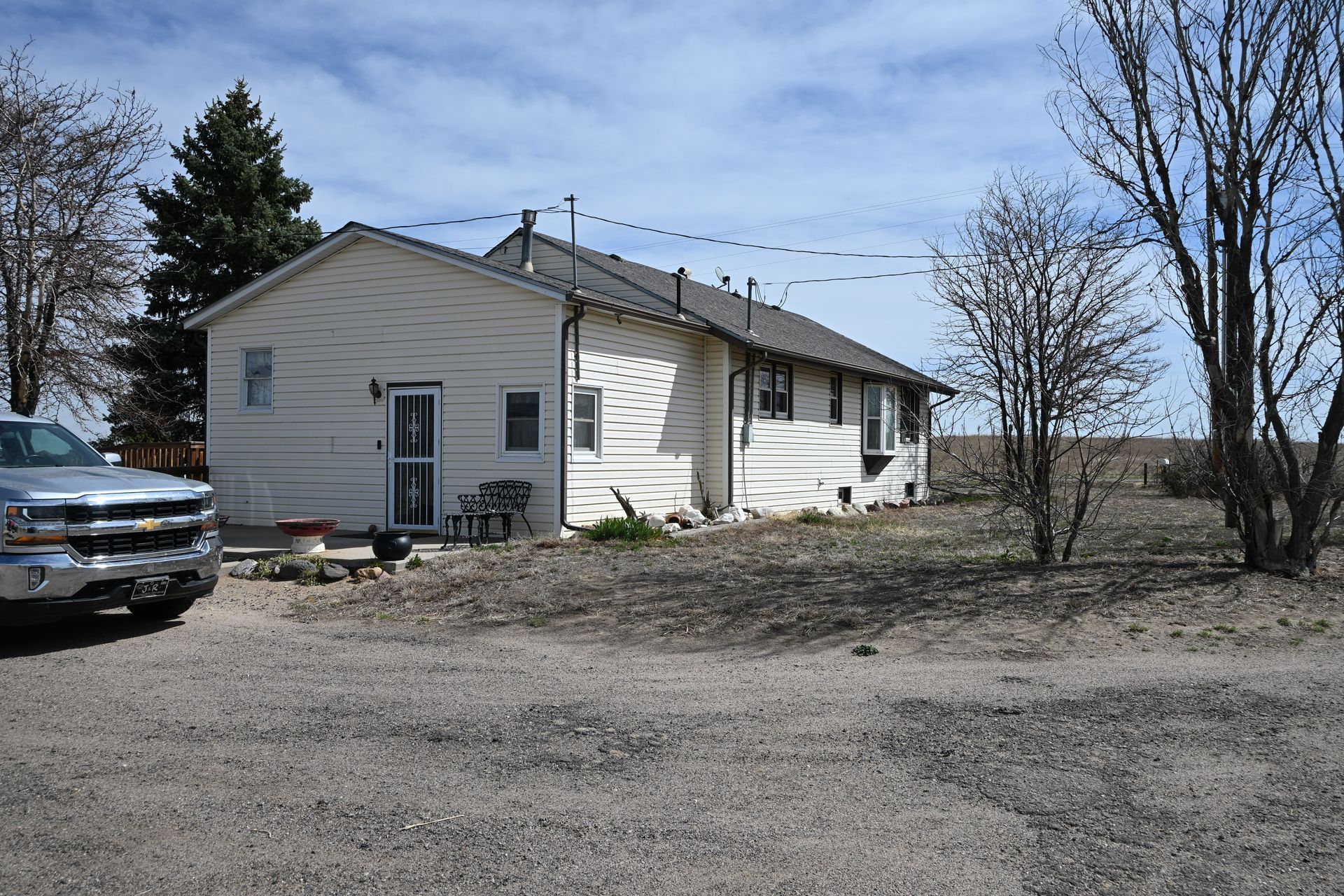 Single-story white house on a gravel lot, with a parked car and bare trees on a sunny day