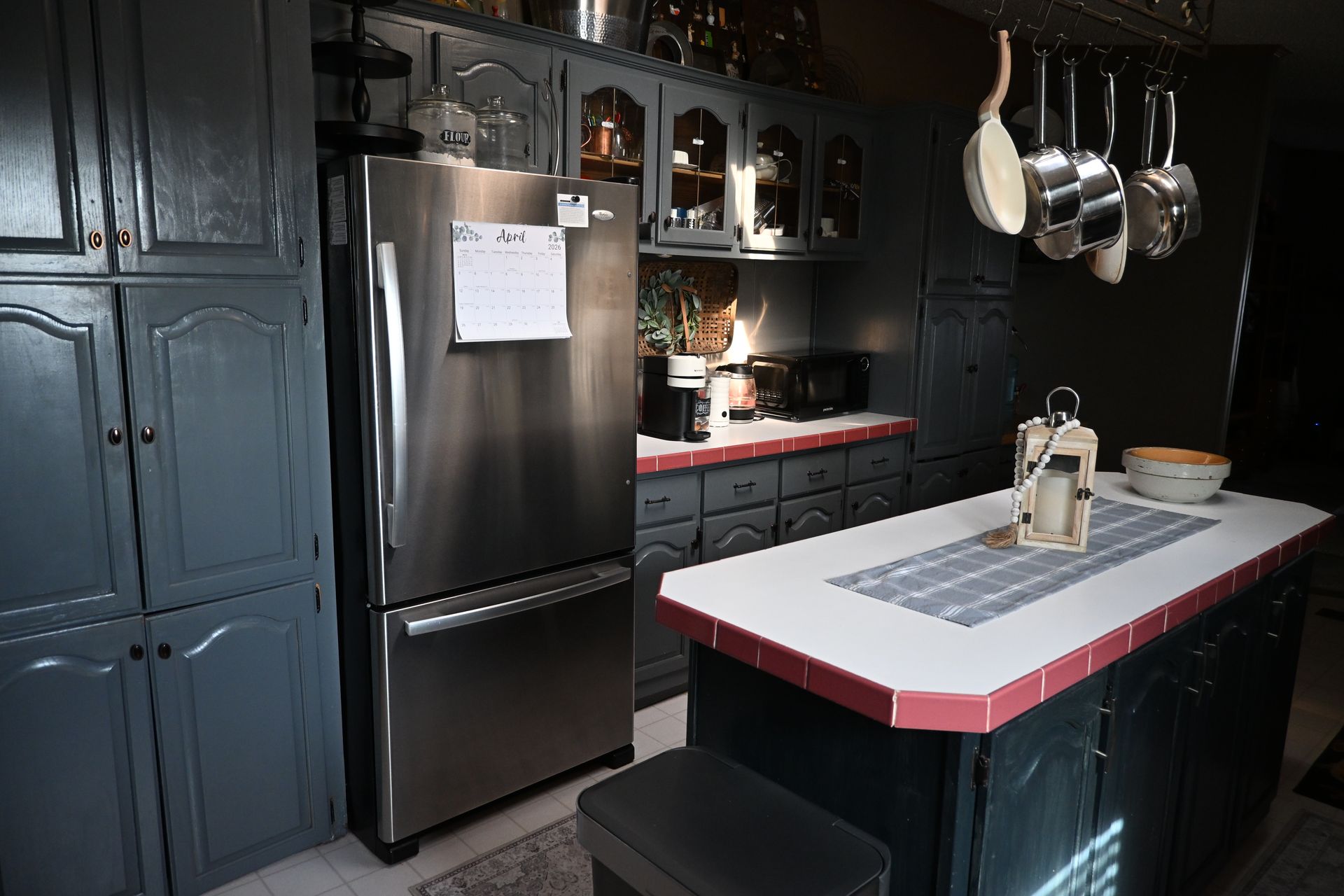 Dark kitchen with stainless fridge, black cabinets, hanging pots, and a white island with red trim.