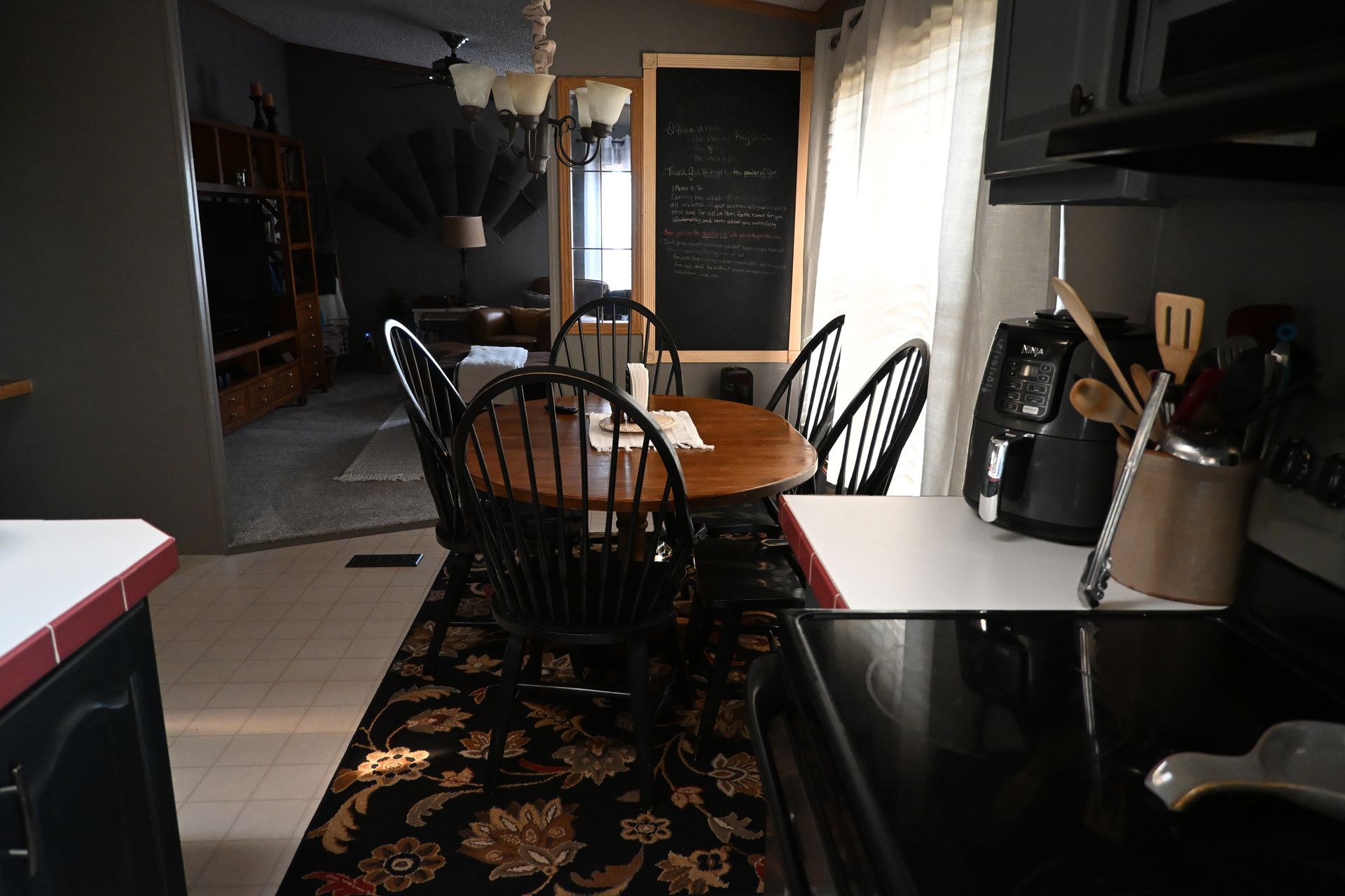 Dim kitchen with a dining table, black chairs, and sunlight through a window.