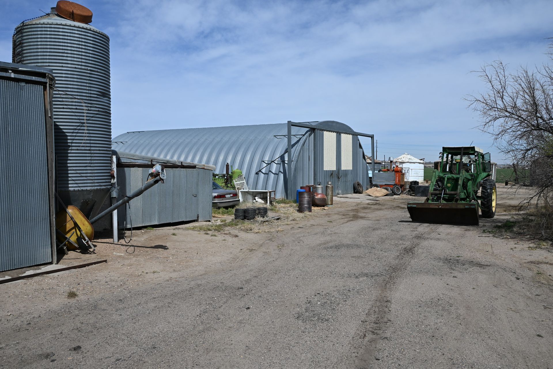 Farmyard with grain silos, metal sheds, and a green tractor on a gravel lane