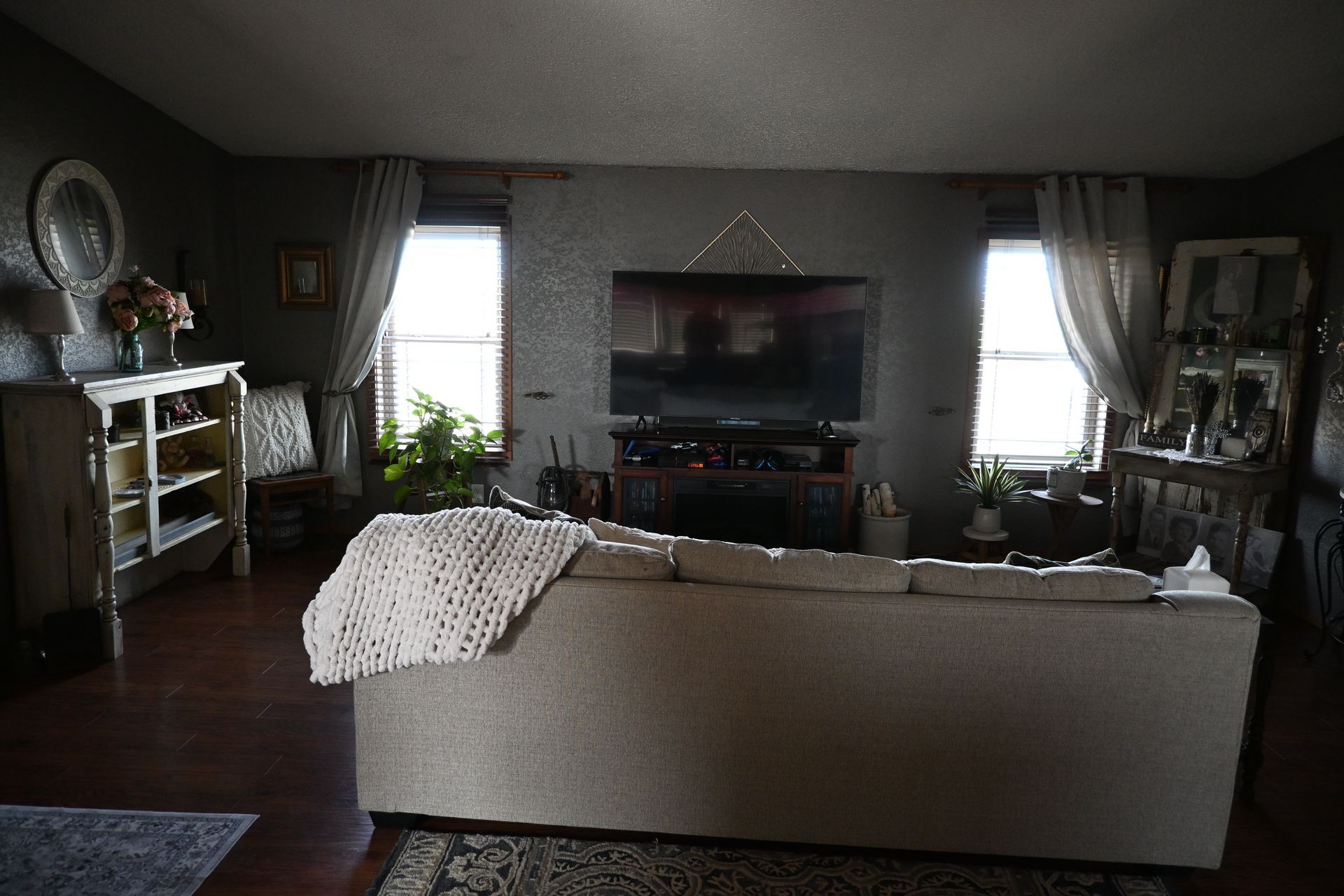 Living room with a beige sofa, TV, windows, and shelves in a dim interior