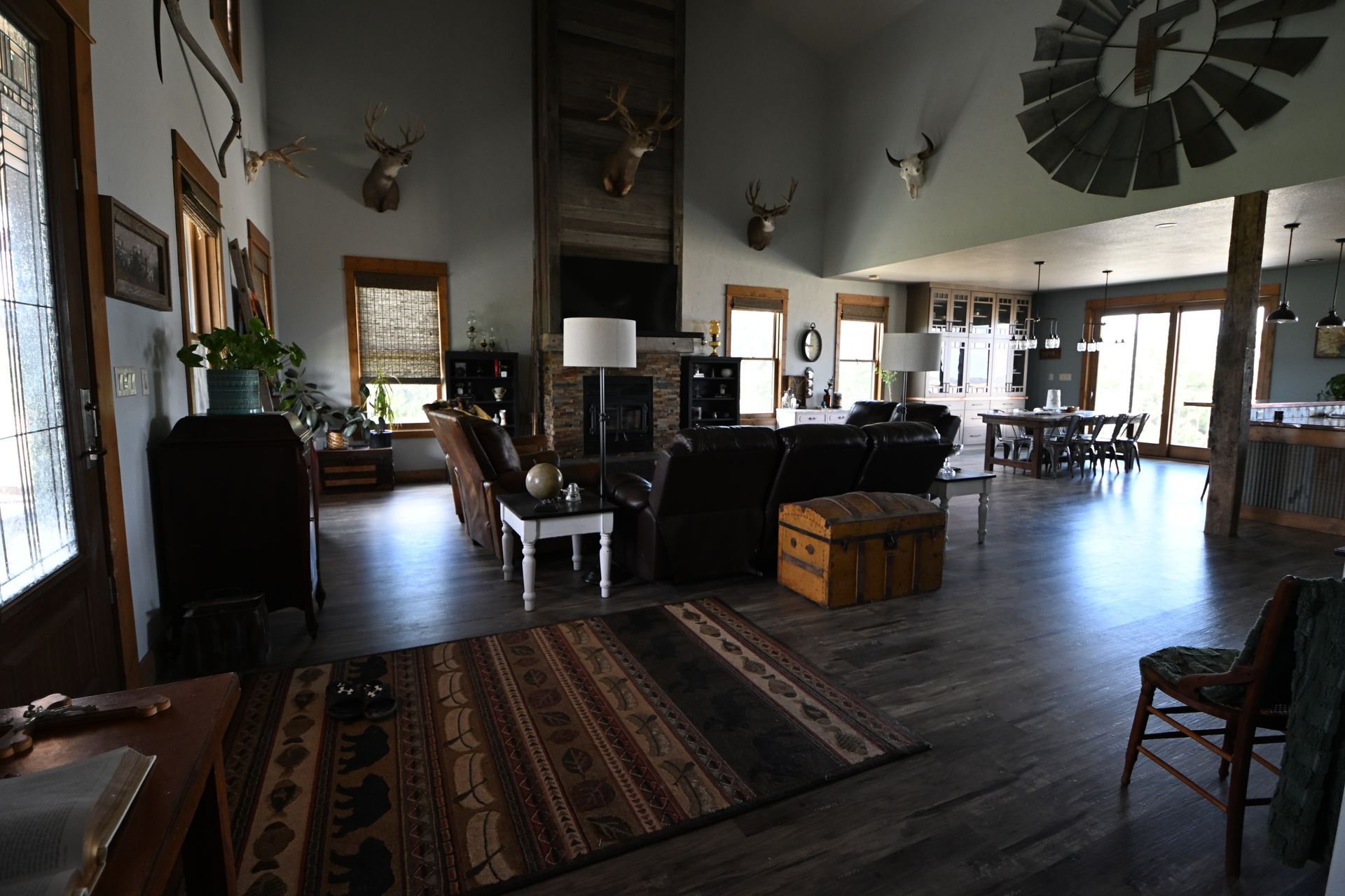 Spacious rustic living room with dark furniture, rugs, mounted decor, and large windows