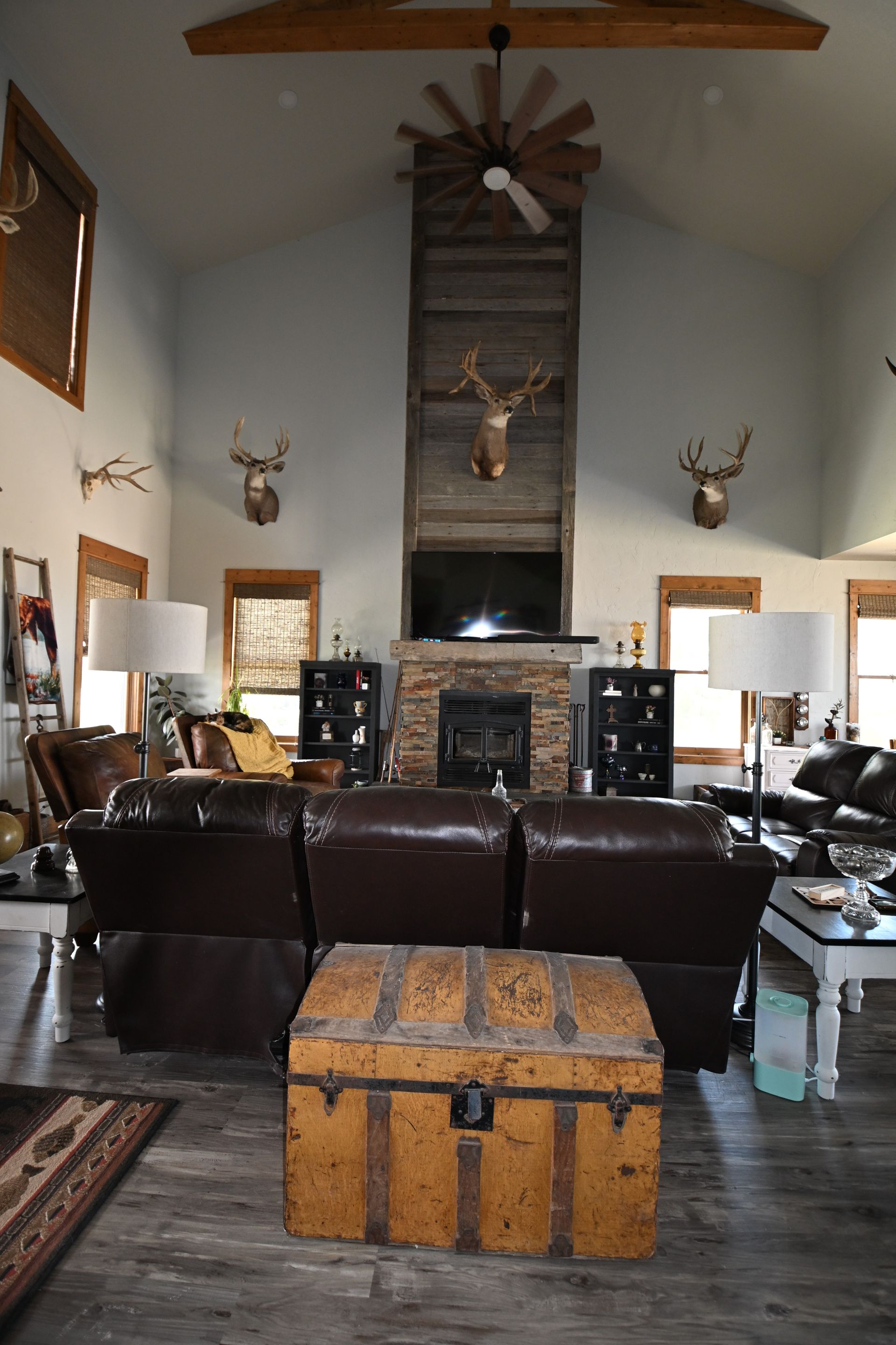 Rustic living room with leather sofa, stone fireplace, mounted antlers, and a wooden chest in the foreground