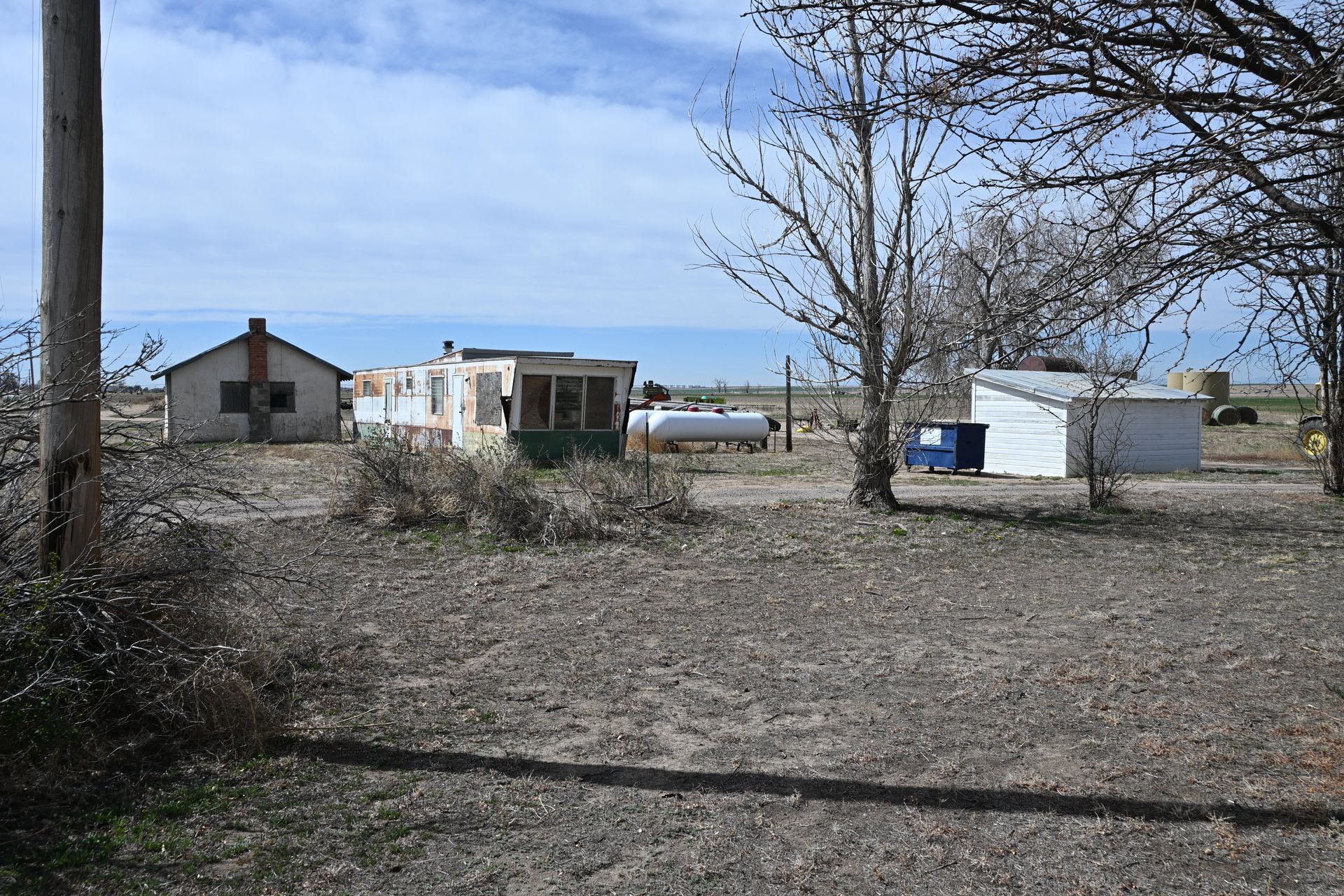 Rural yard with small buildings, leafless trees, and an open dirt lot under a blue sky