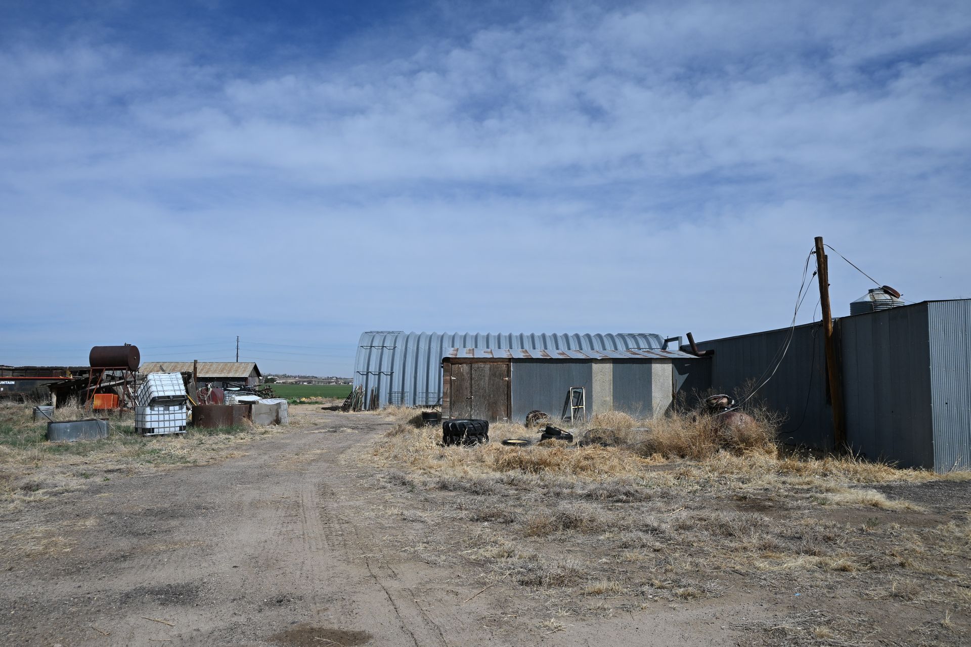 Rural gravel yard with metal sheds, dry grass, and a cloudy blue sky