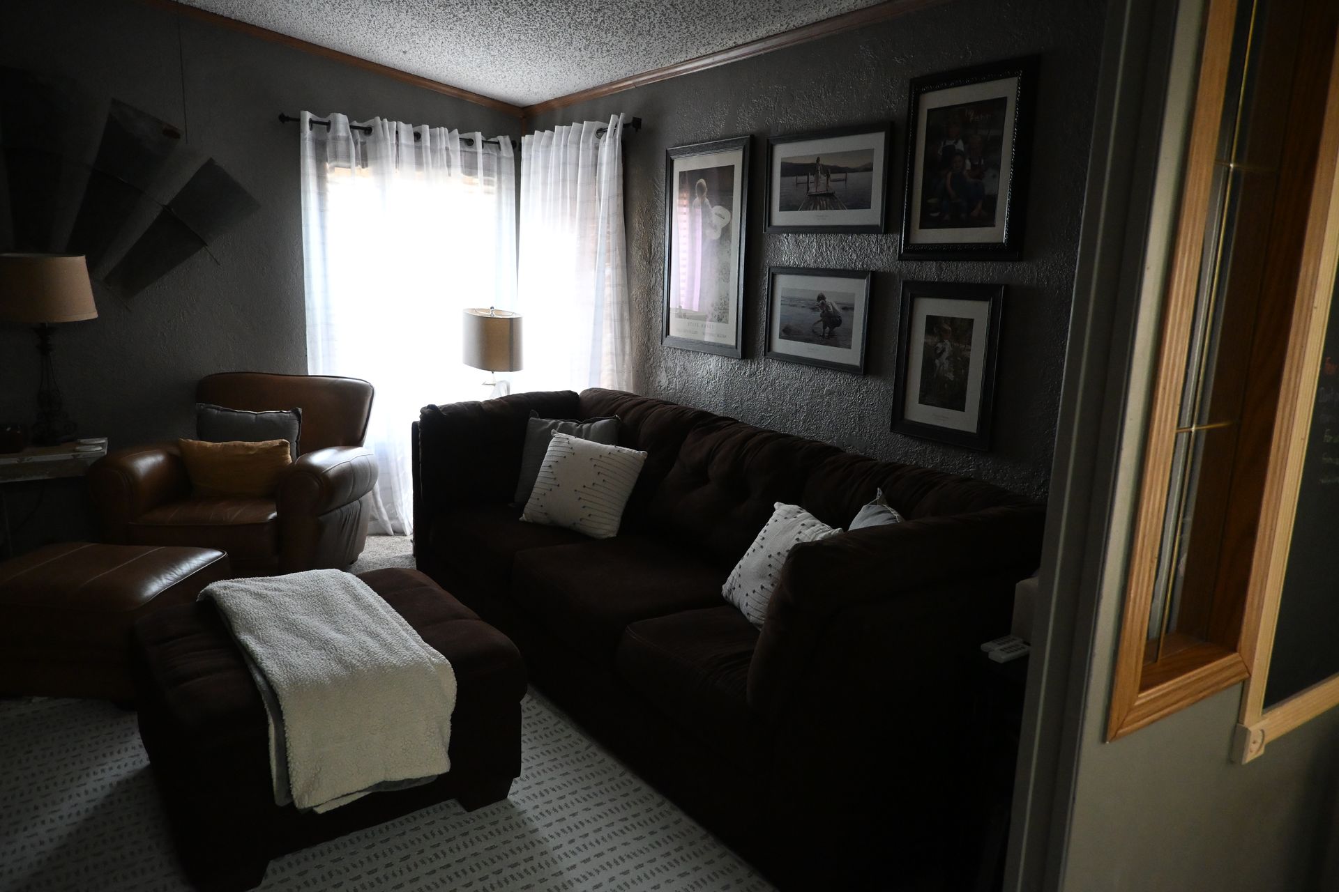 Dim living room with dark sofa, armchair, ottoman, framed wall art, and bright window curtains
